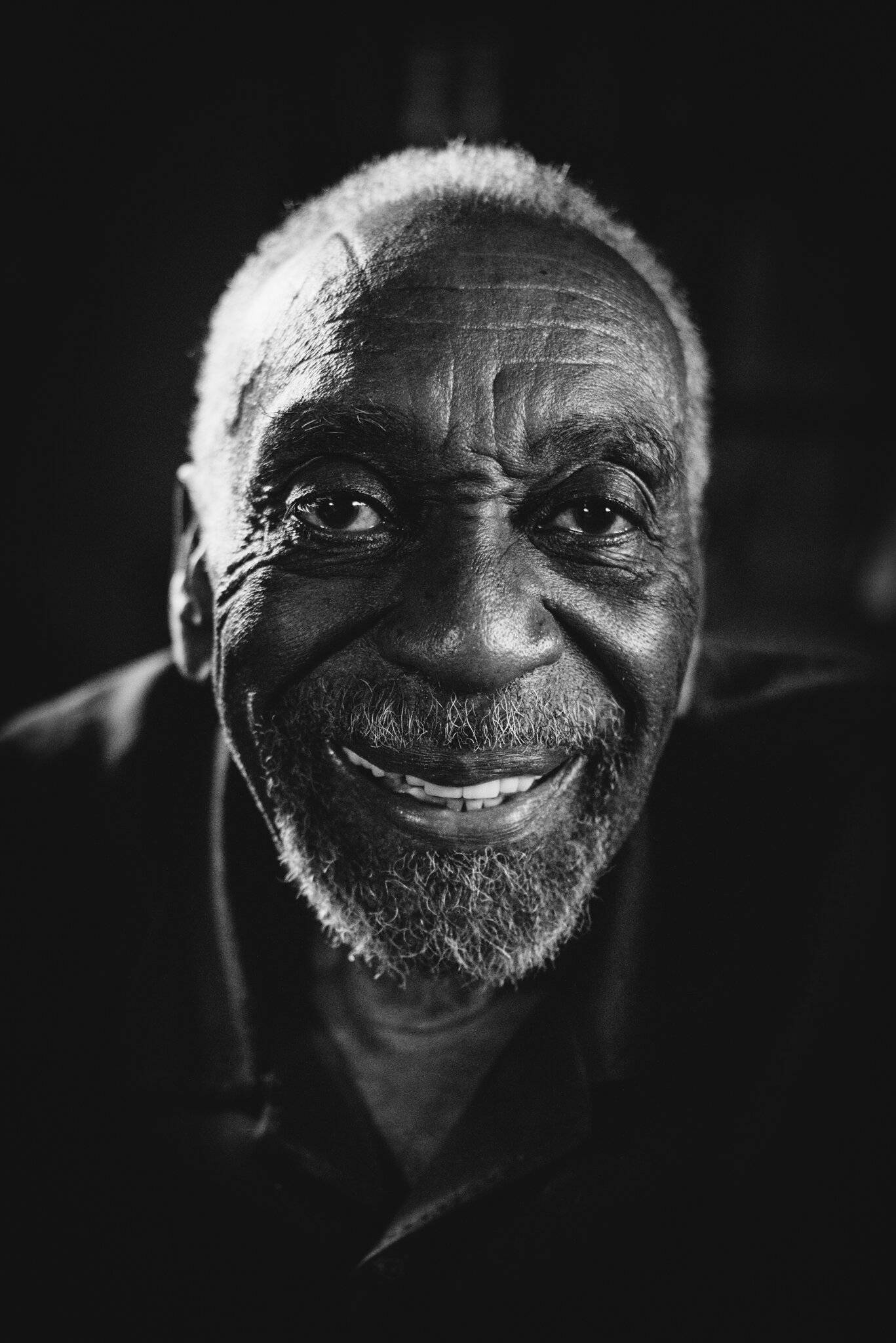 actor 桌布 壁紙 — 一個特寫黑白肖像，一位年長的黑人男性，帶著溫暖的笑容和深深的皺紋，背景為深色。