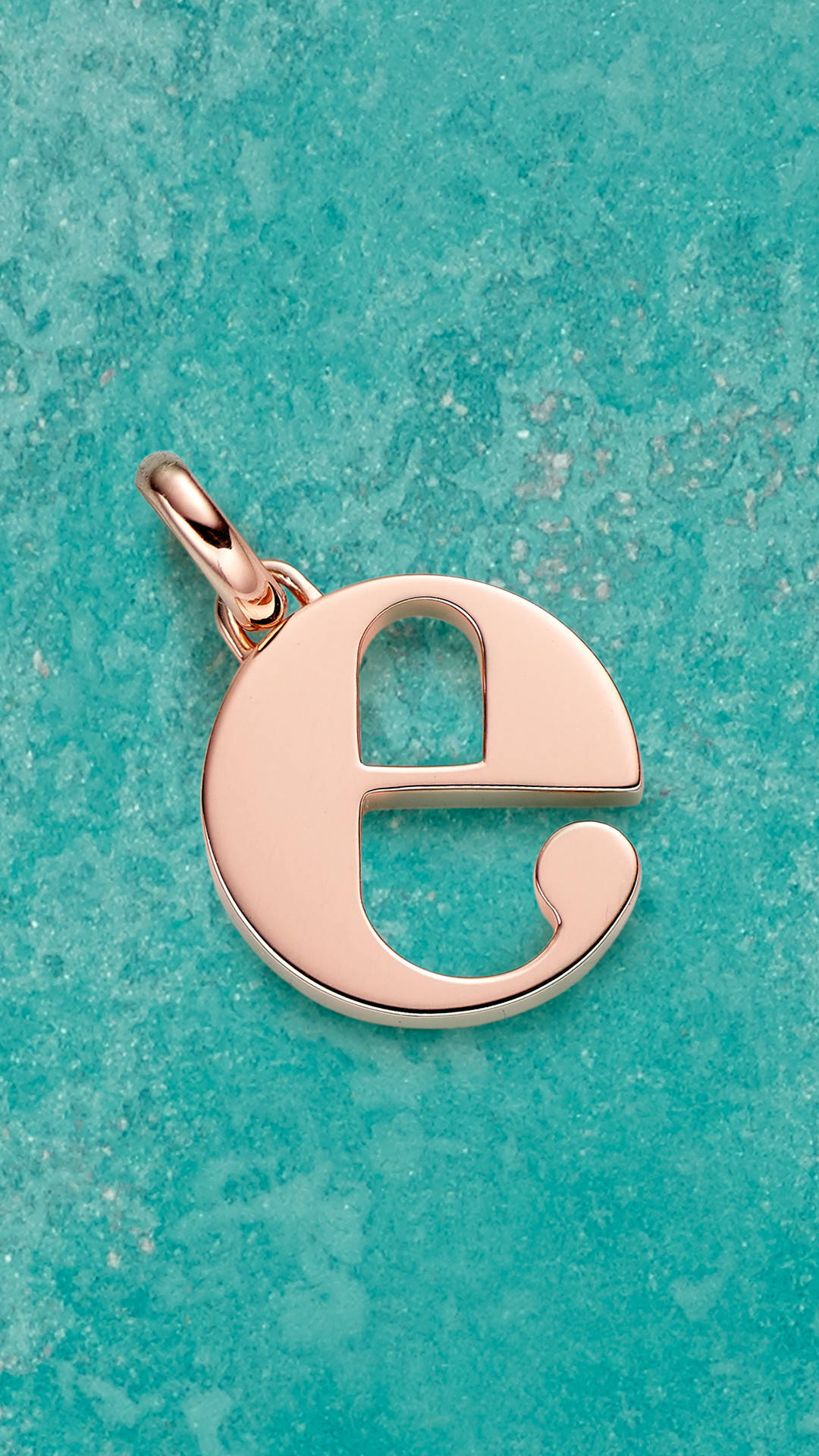 E 桌布 壁紙 — 一個玫瑰金字母e吊墜放置在帶有柔和光線的紋理青綠色表面上。