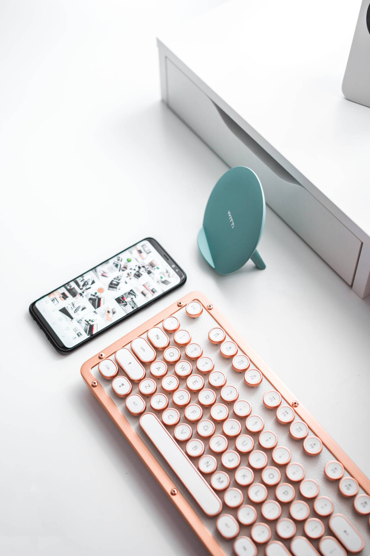 iPhone 桌布 壁紙 — 一個玫瑰金復古鍵盤放在一支智慧型手機和一個青綠色無線充電器旁邊，置於簡約白色桌上。