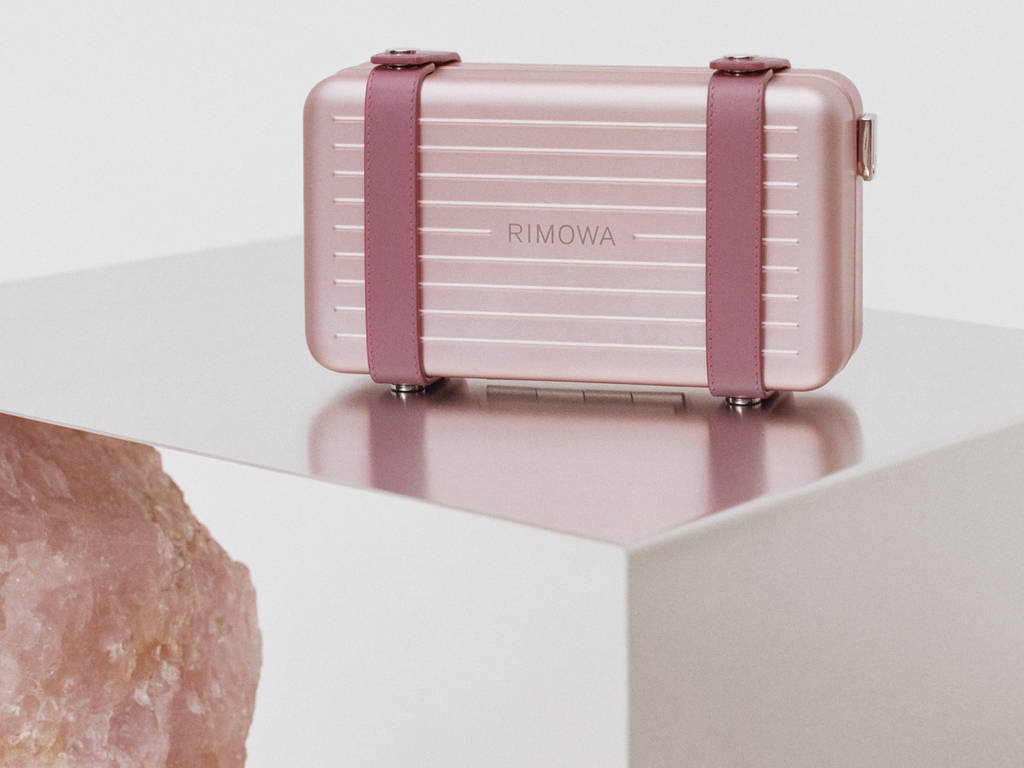 時尚 桌布 壁紙 — 一個玫瑰金色Rimowa行李箱配紫紅色帶子，優雅地放在白色底座上，旁邊有一個粉紅色水晶。