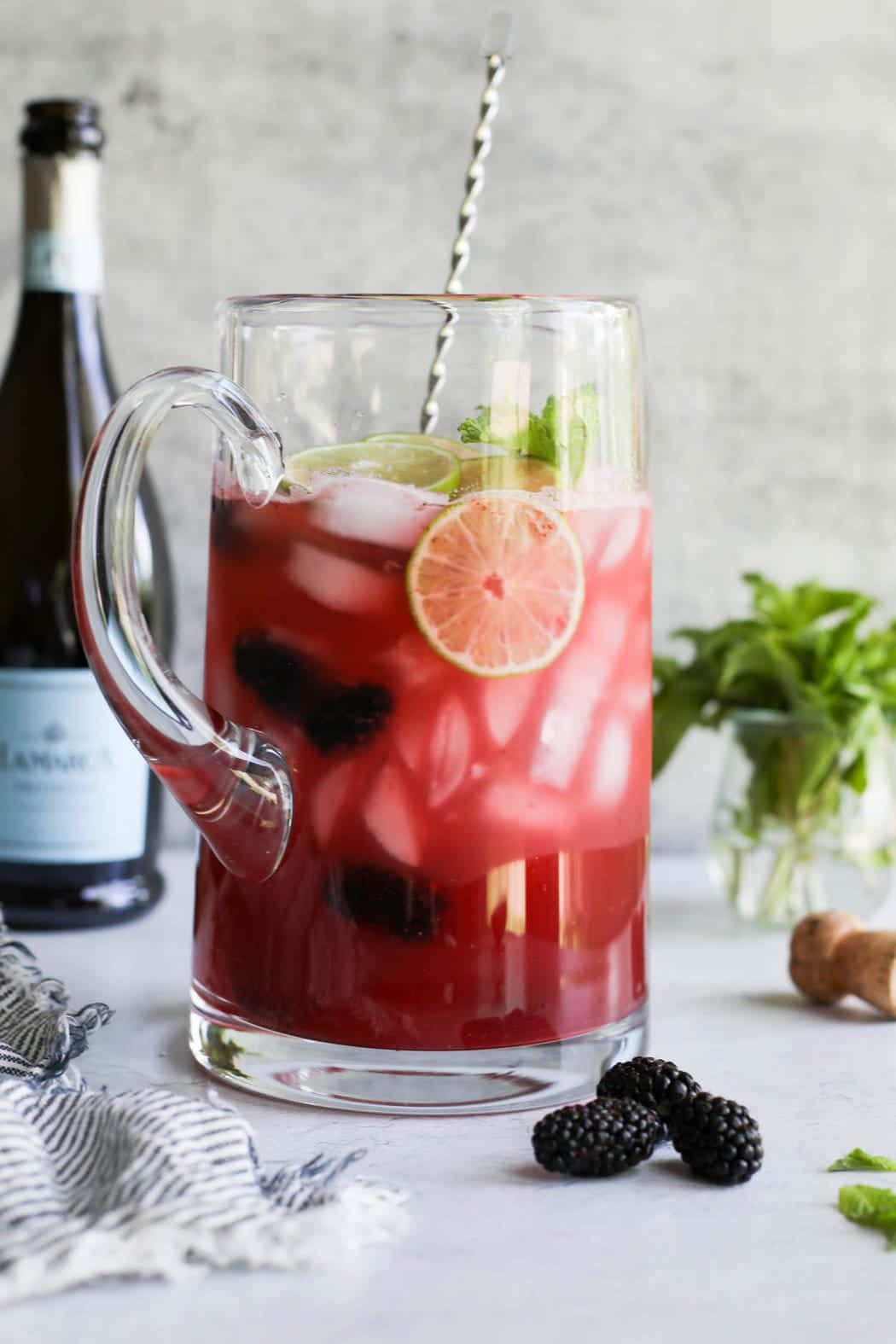 Beautiful Drink 桌布 壁紙 — 一個玻璃壺中裝有充滿活力的紅色莓果飲料，飾以青檸、薄荷和冰塊，放在白色表面上，旁邊是新鮮黑莓和一瓶葡萄酒。