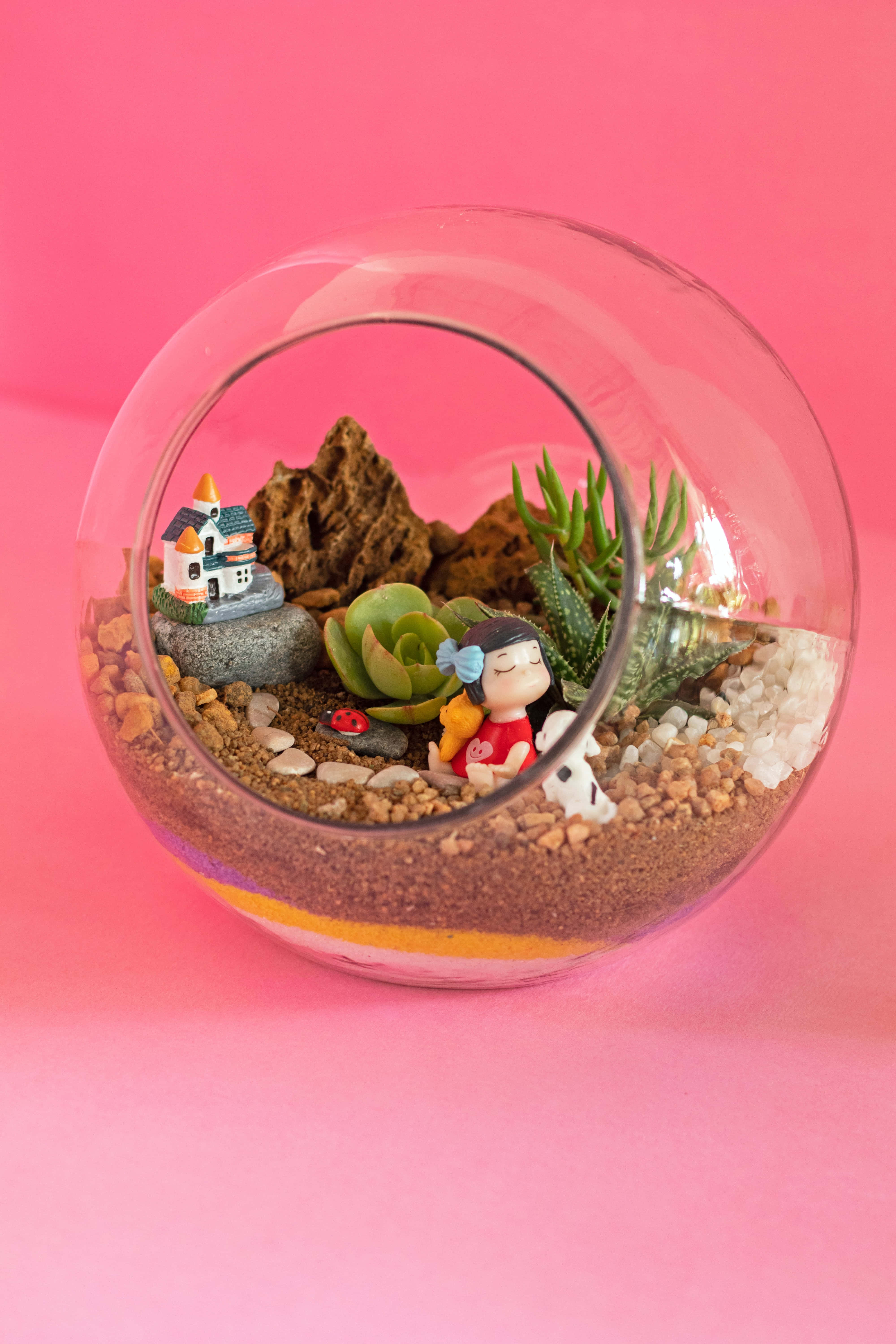 Terrarium 桌布 壁紙 — 一個玻璃容器內有一個微型花園，裡面有肉質植物、石頭、一間小房子和一個在粉紅色背景上靜靜休息的人偶。