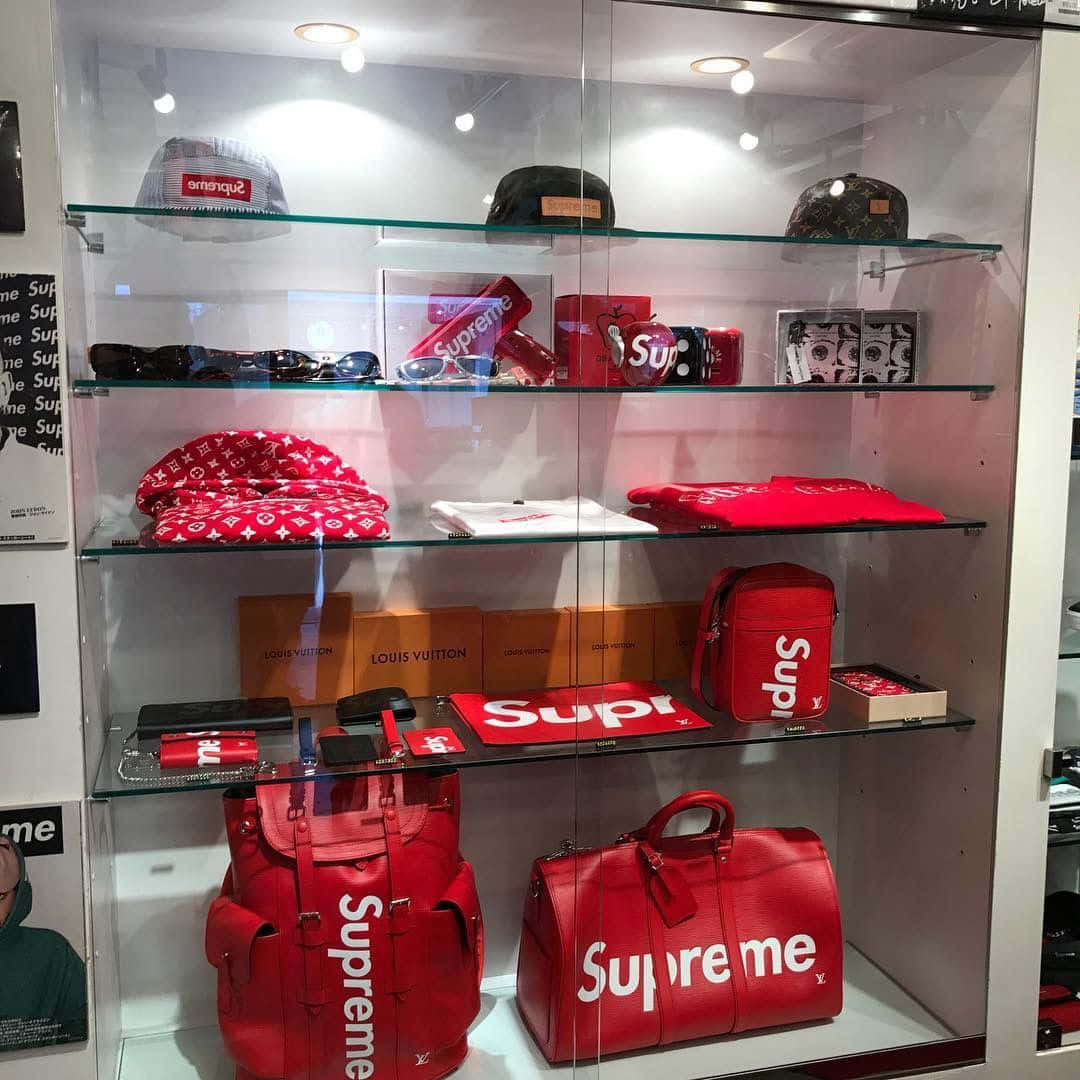 supreme 桌布 壁紙 — 一個玻璃展示櫃，裝滿了紅色Supreme品牌物品，包括手提包、帽子和配件，以及Louis Vuitton盒子，在明亮的燈光下。