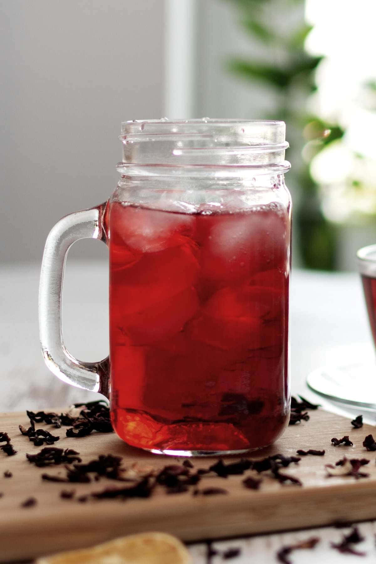 Beautiful Drink 桌布 壁紙 — 一個玻璃梅森罐裝滿了充滿活力的紅色木槿冰茶和冰塊，放在一個散落著乾燥木槿花瓣的木板上。