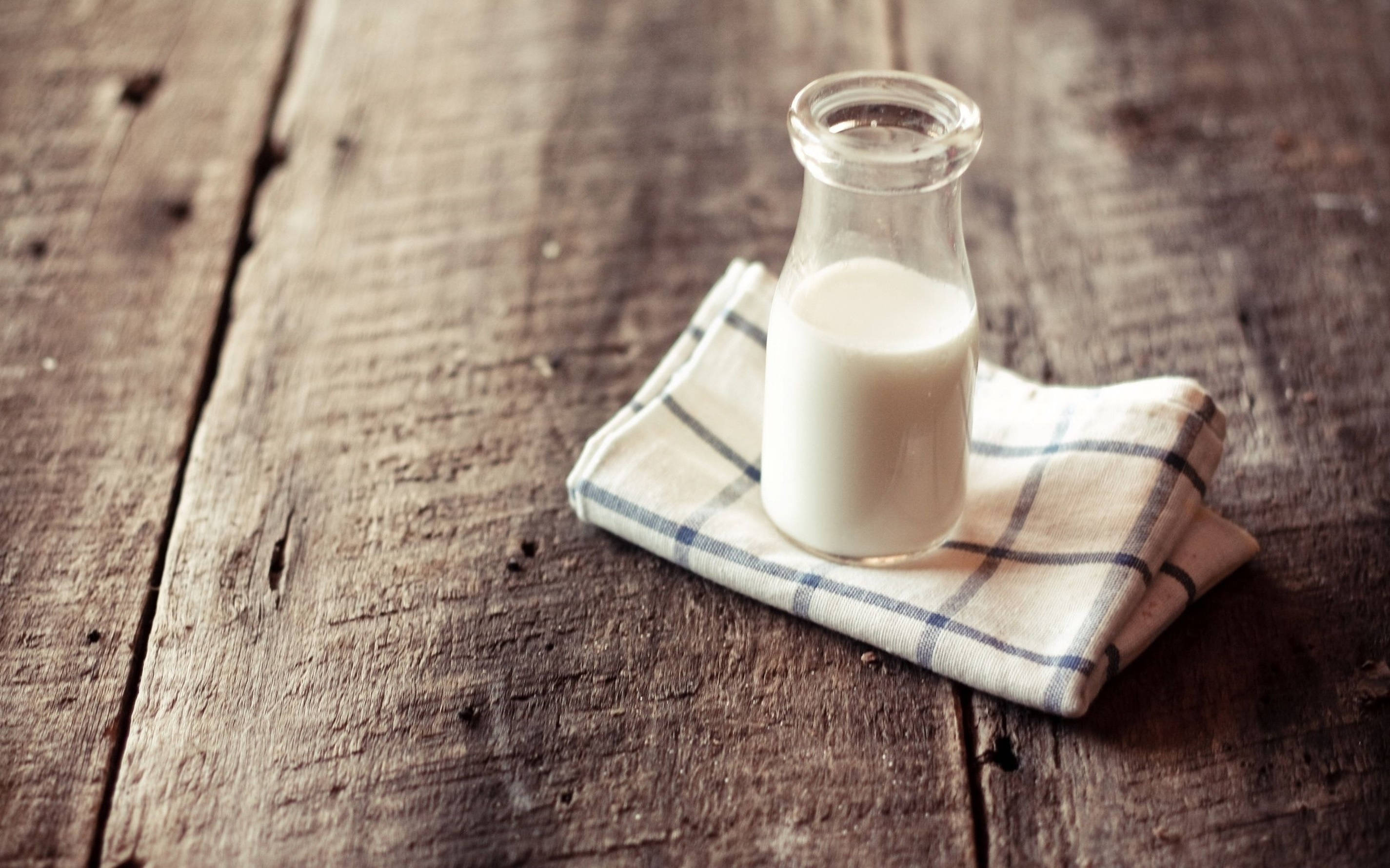Milk 桌布 壁紙 — 一個玻璃牛奶瓶放在一張磨損的木桌上，桌上鋪著格子布，帶有溫暖質樸的燈光。