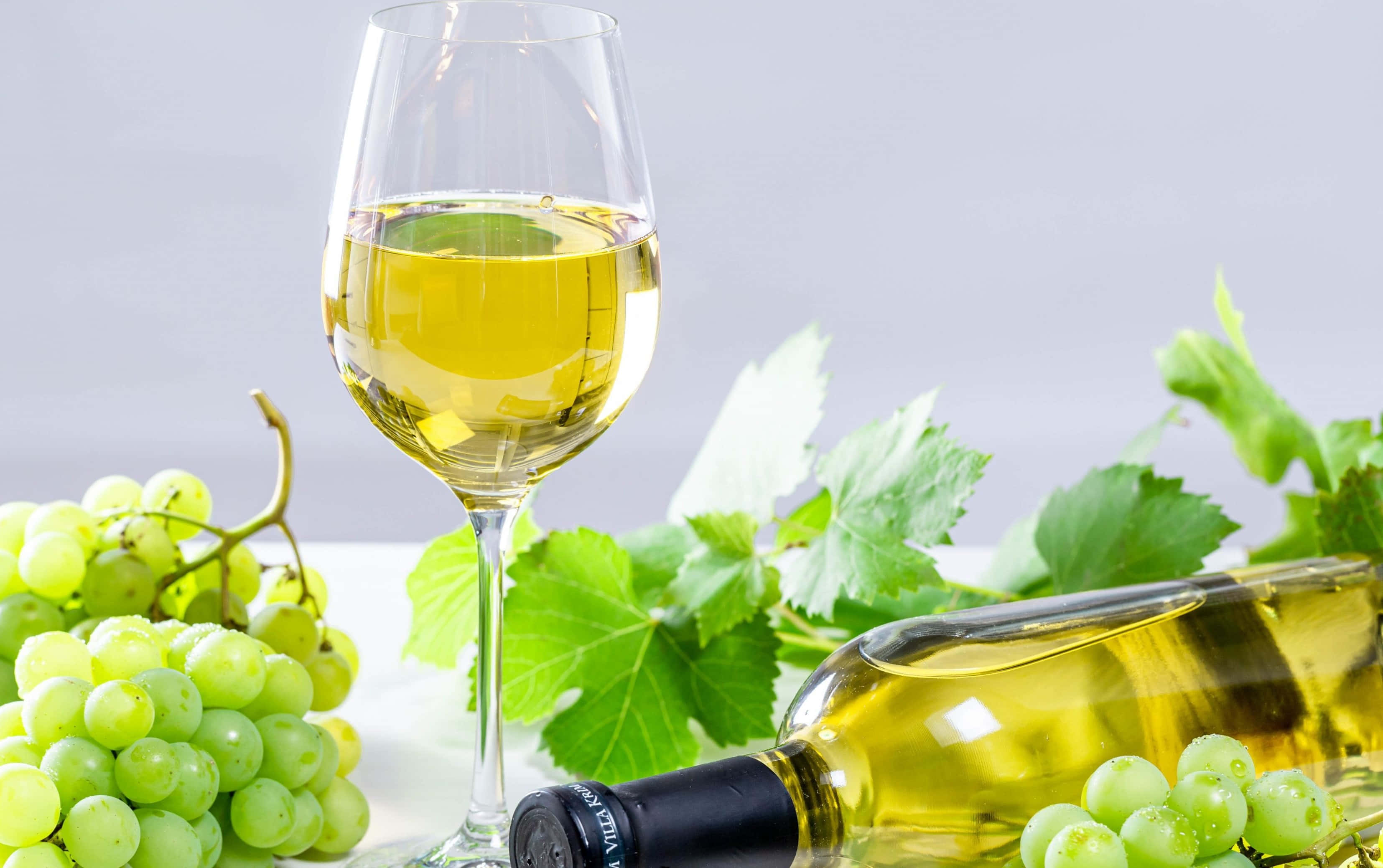 White Wine 桌布 壁紙 — 一個玻璃瓶和裝滿酒的酒杯，周圍有青葡萄和葉子，放在潔白的表面上。