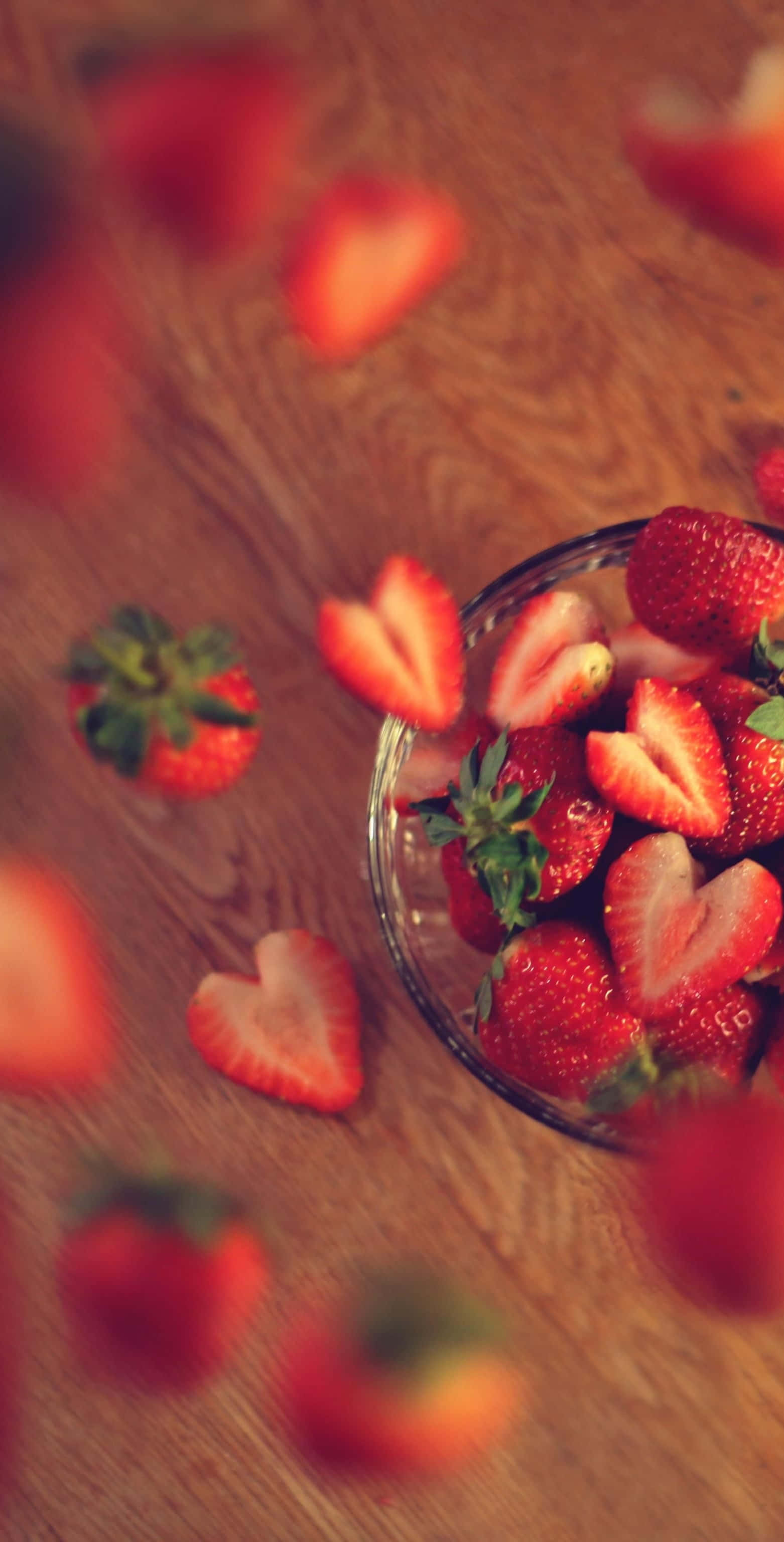Cute Strawberry 桌布 壁紙 — 一個玻璃碗裝滿新鮮草莓，有些切成心形，放在溫暖的木桌上，周圍散落著水果。
