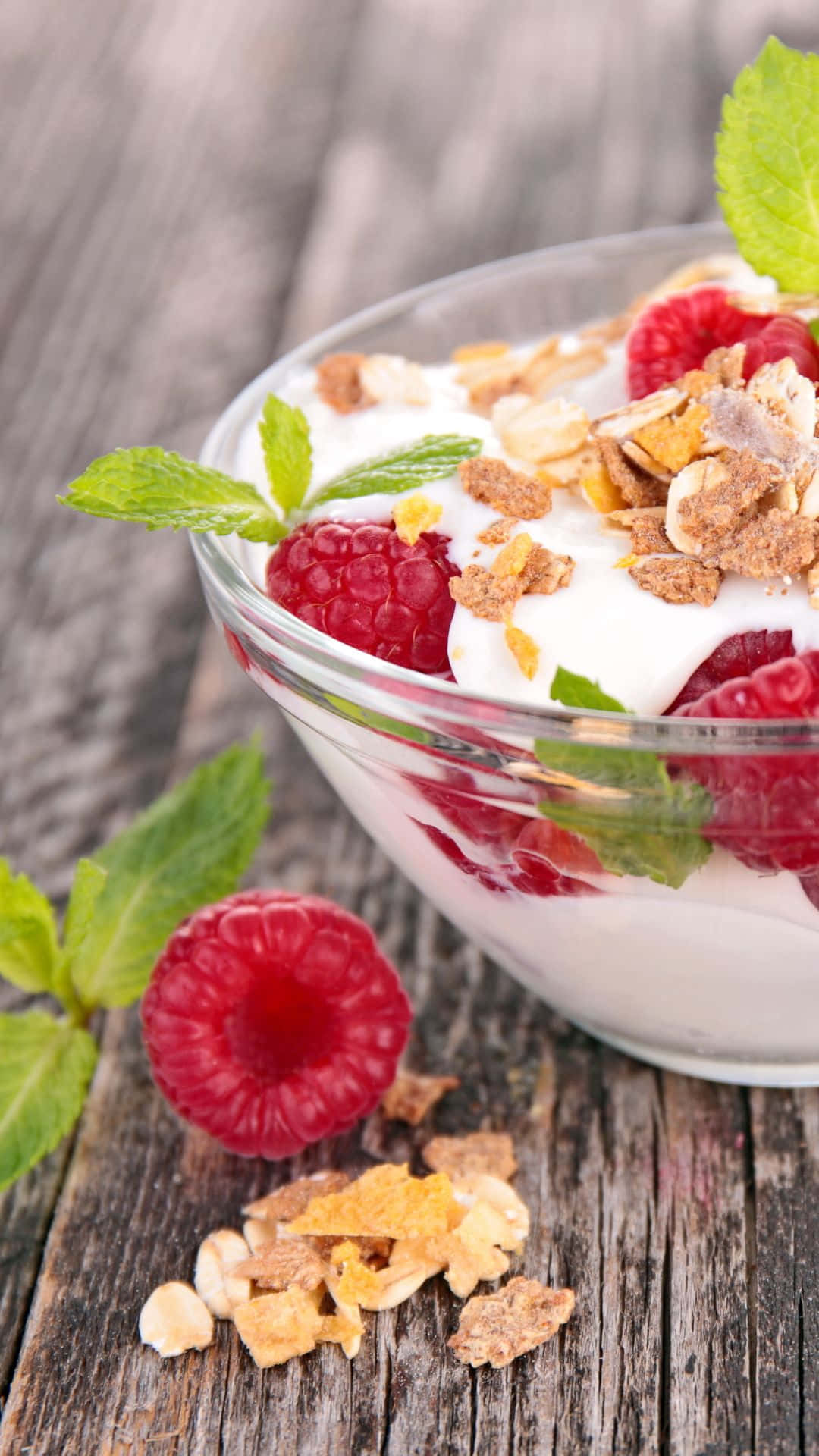 Yogurt 桌布 壁紙 — 一個玻璃碗裝著奶油狀優格，上面放上新鮮覆盆子、格蘭諾拉麥片和薄荷葉，放在質樸的木質表面上。