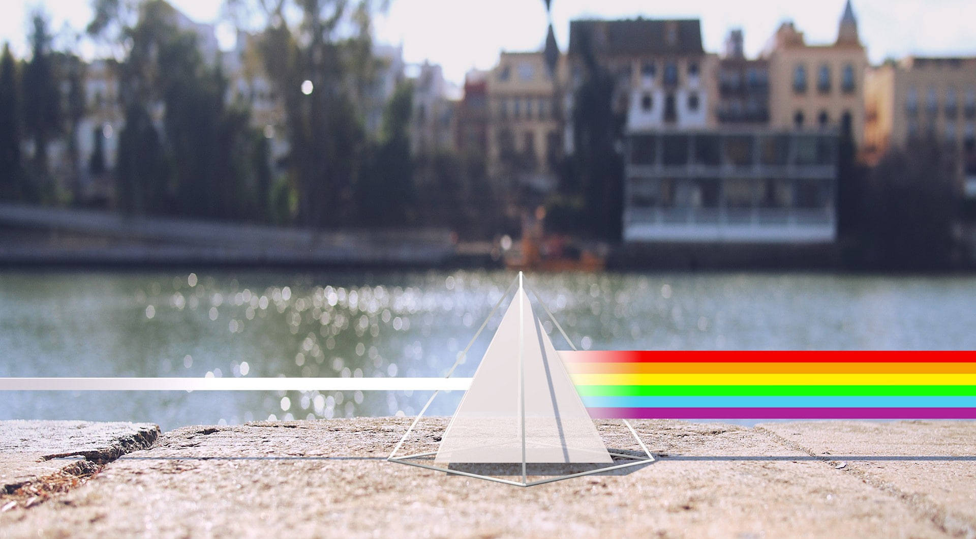 Prism 桌布 壁紙 — 一個玻璃稜鏡放在石架上，將陽光折射成一道鮮豔的彩虹，橫跨河流，背景是模糊的城市建築。