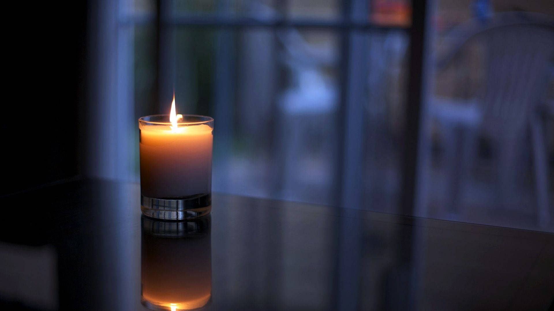 蠟燭 桌布 壁紙 — 一個玻璃罐中的單支點燃蠟燭在反射表面上投射出溫暖的光芒，背後是模糊的窗景。