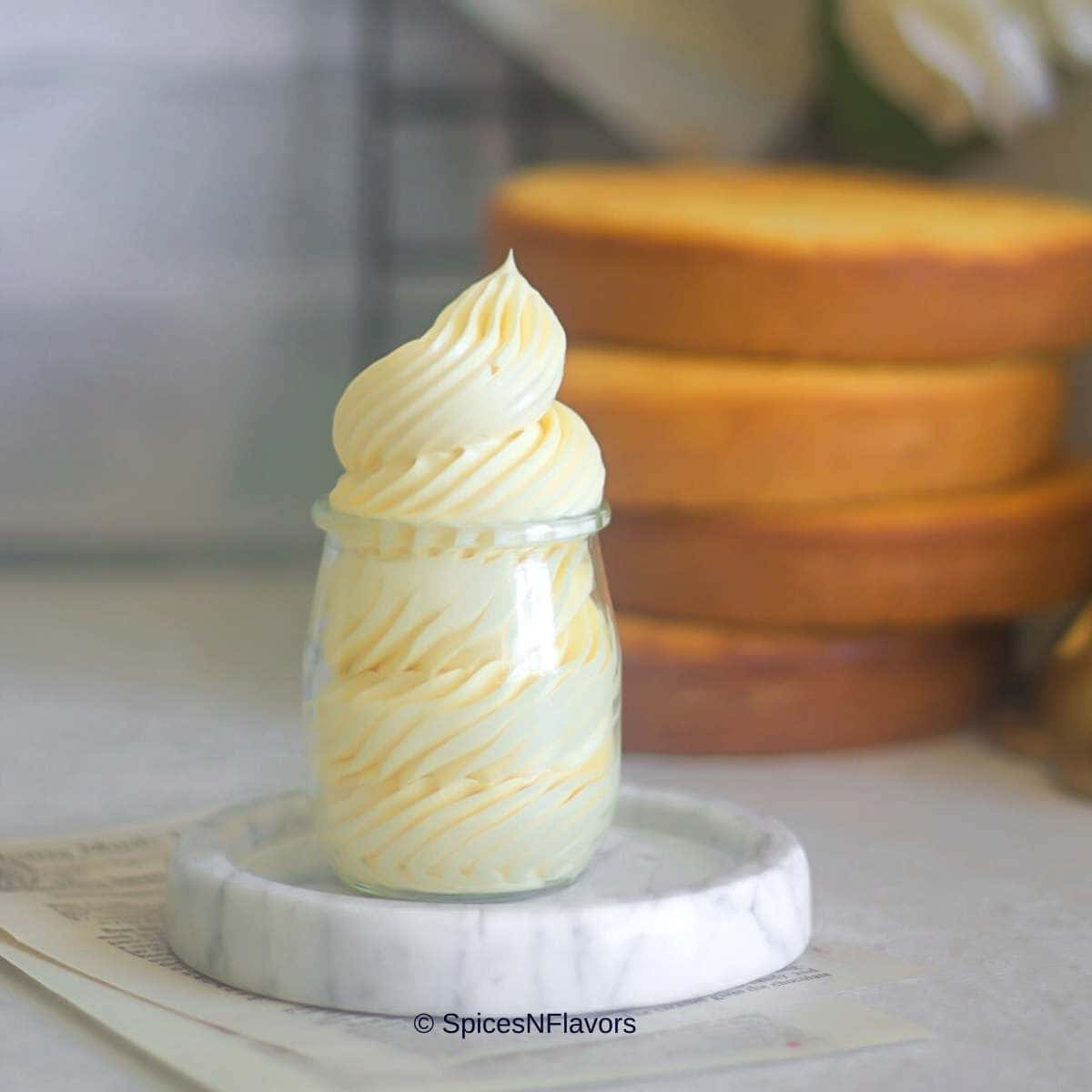 Buttercream 桌布 壁紙 — 一個玻璃罐裝滿了奶油香草奶油糖霜，放在大理石杯墊上，背景有模糊的蛋糕層。
