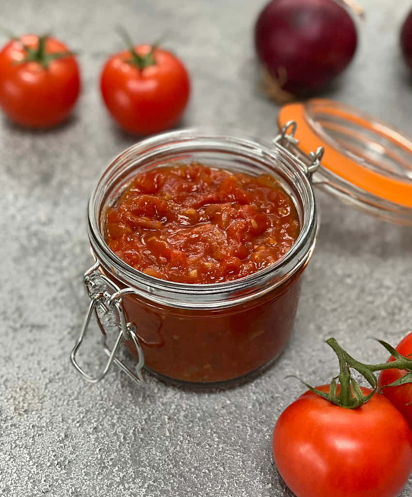 Red Sauce 桌布 壁紙 — 一個玻璃罐裝滿了粗粒的紅色番茄醬，放在灰色表面上，周圍是新鮮番茄和洋蔥。