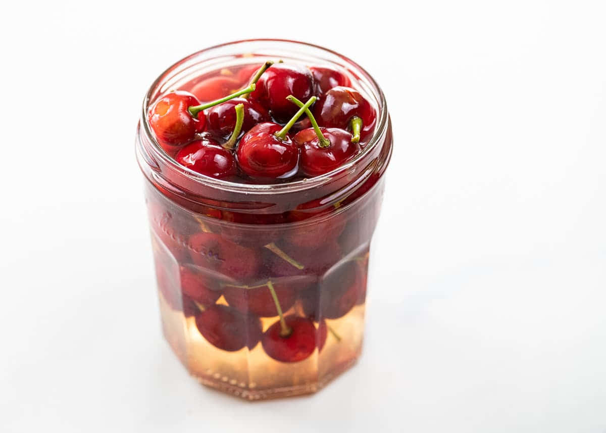 Red Cherries 桌布 壁紙 — 一個玻璃罐裝滿光澤紅櫻桃，浸泡在琥珀色液體中，背景為純白。