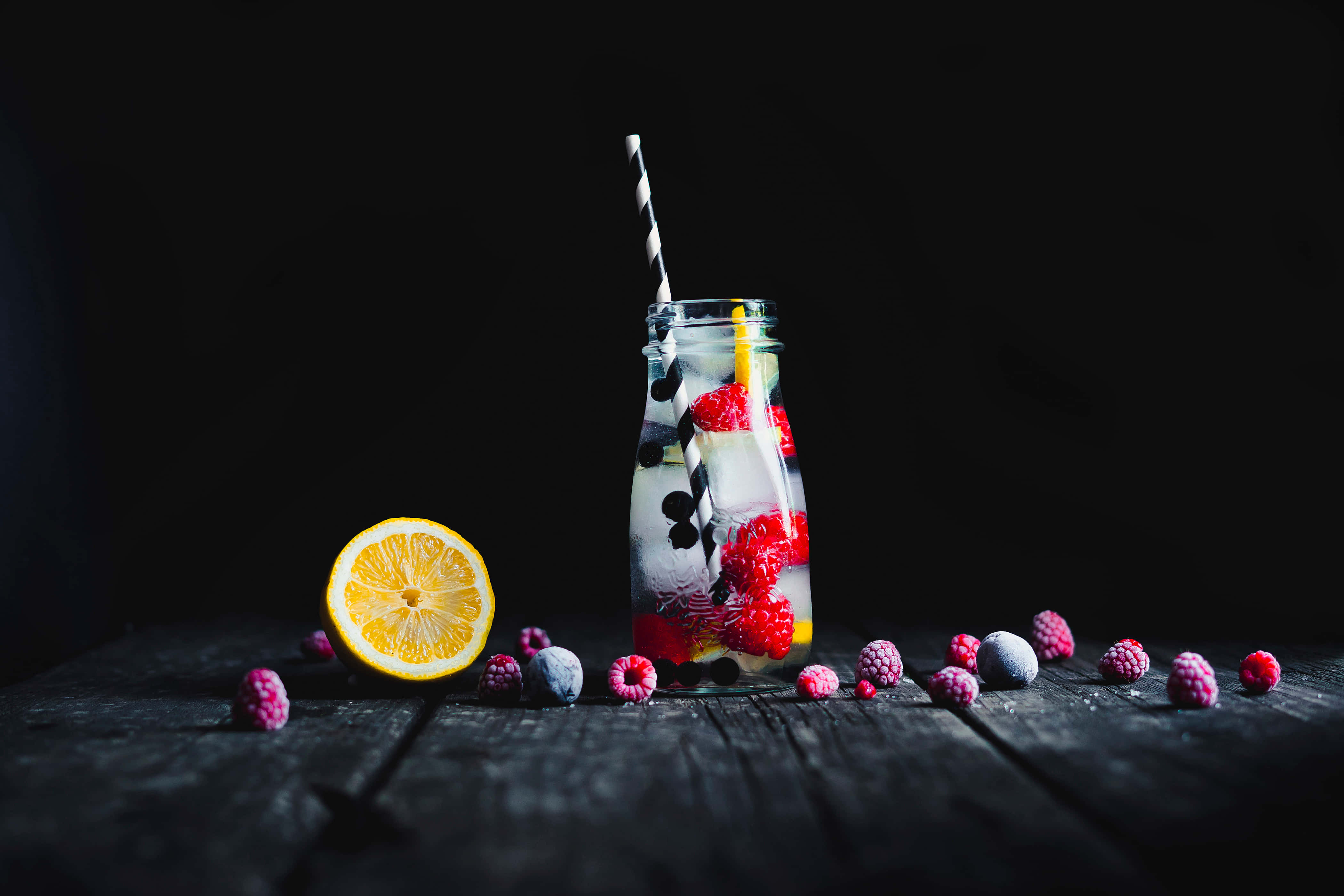 Food And Beverage 桌布 壁紙 — 一個玻璃罐裝滿冰莓和檸檬水，放在深色木桌上，周圍散落著水果，背景為黑色。