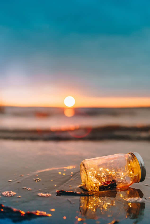 海灘 桌布 壁紙 — 一個玻璃罐裝滿發光的仙女燈，放在日落時分的濕沙上，映照出金色溫暖，襯托柔和的藍天。