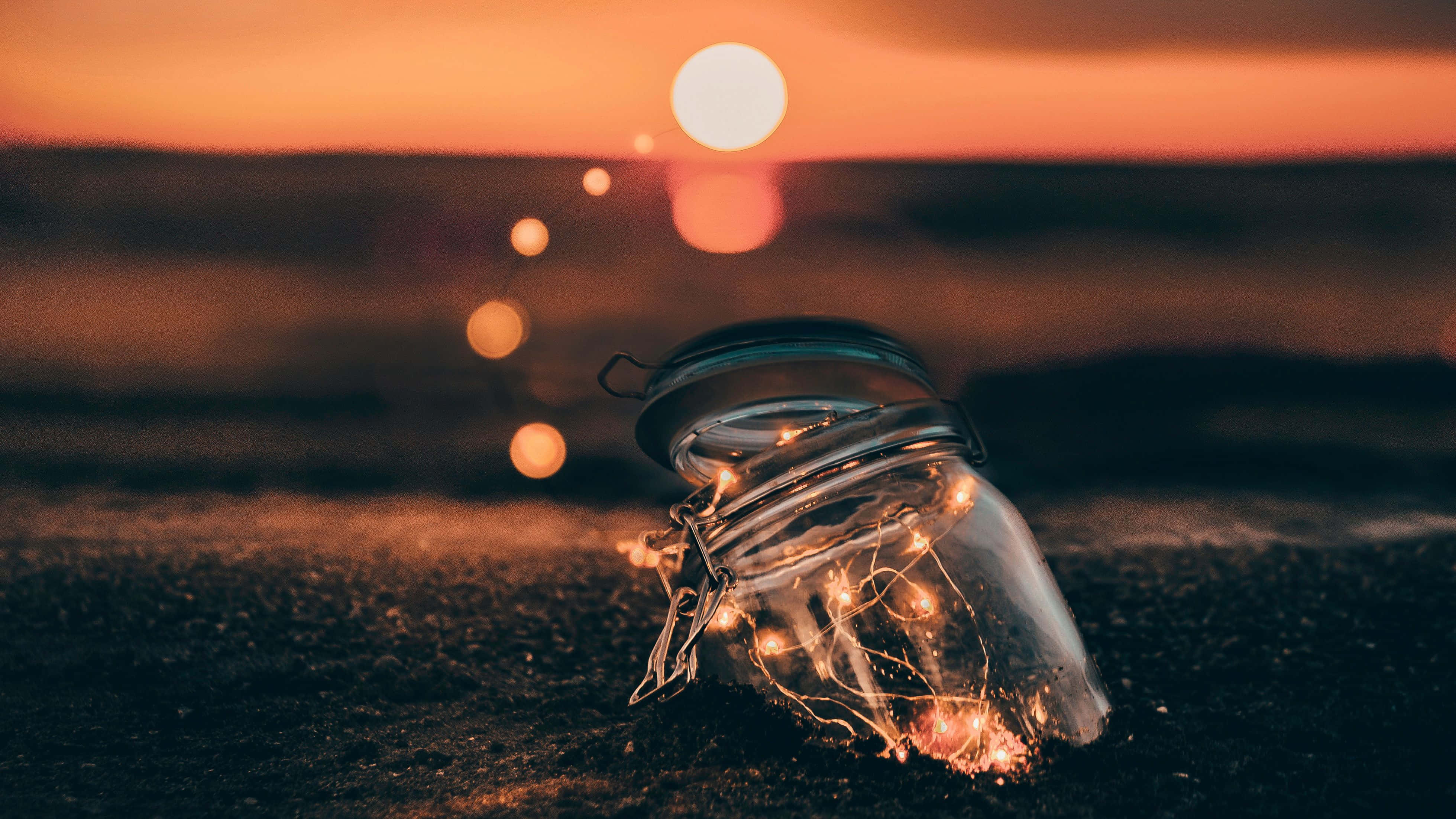 Light Effect 桌布 壁紙 — 一個玻璃罐裝滿發光的仙女燈，放在深色沙灘上，夕陽下背景有溫暖的散景燈光。