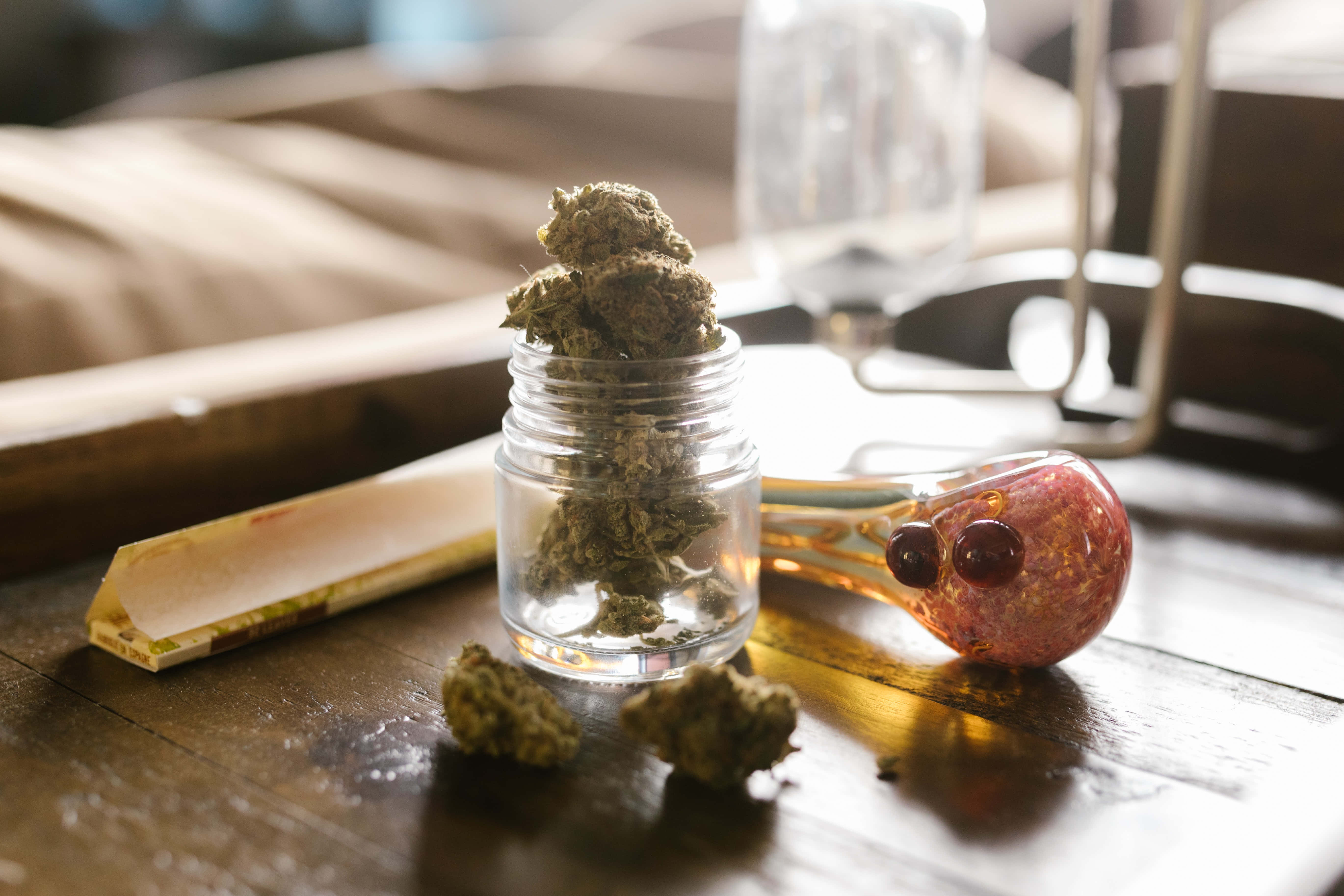 Weed Blunt 桌布 壁紙 — 一個玻璃罐裝滿綠色大麻花蕾，放在木桌上，旁邊有一個紅色玻璃水煙槍和捲菸紙。