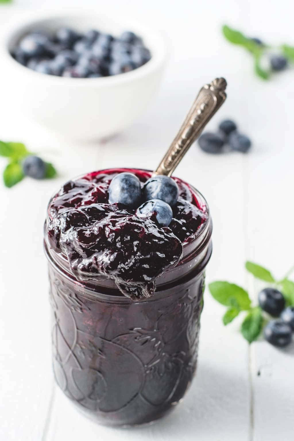 Blueberry Jam 桌布 壁紙 — 一個玻璃罐裝滿閃亮的藍莓果醬，上面放上新鮮莓果和復古湯匙，放在明亮的白色木質表面上。