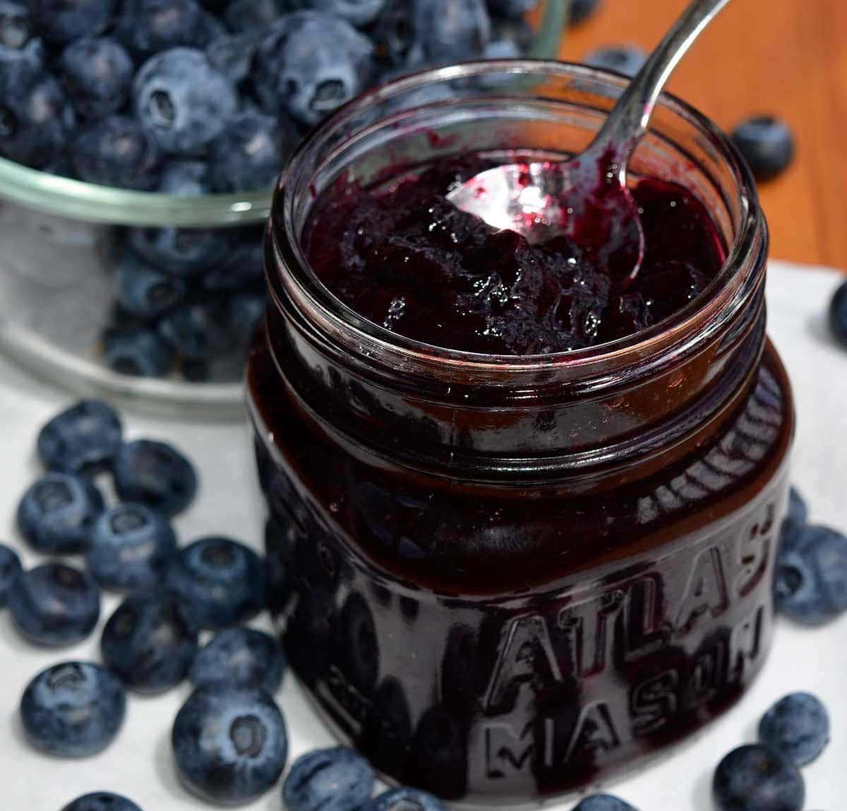Blueberry Jam 桌布 壁紙 — 一個玻璃罐裝著濃郁深藍色的藍莓果醬，裡面放著一隻湯匙，周圍是新鮮藍莓。