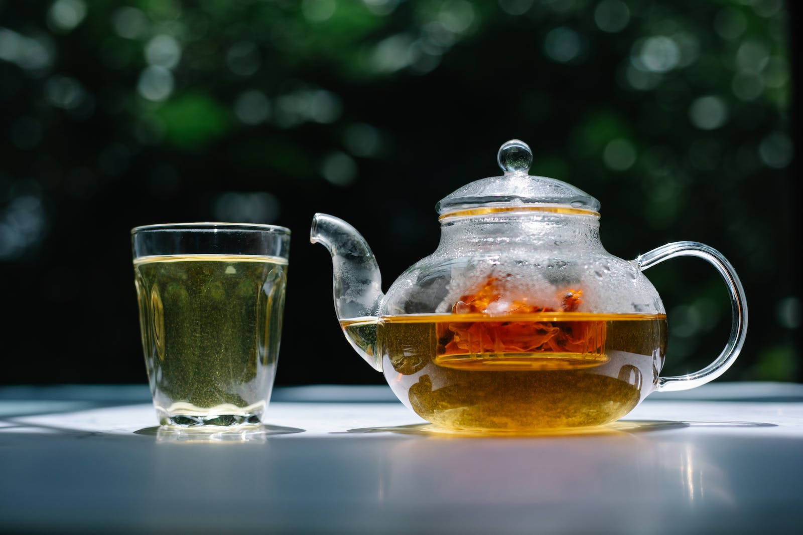 Transparent 桌布 壁紙 — 一個玻璃茶壺，裡面有琥珀色茶和漂浮的花朵，放在一張白色桌子上，旁邊有一個裝滿茶的玻璃杯，背景是模糊的綠色花園。