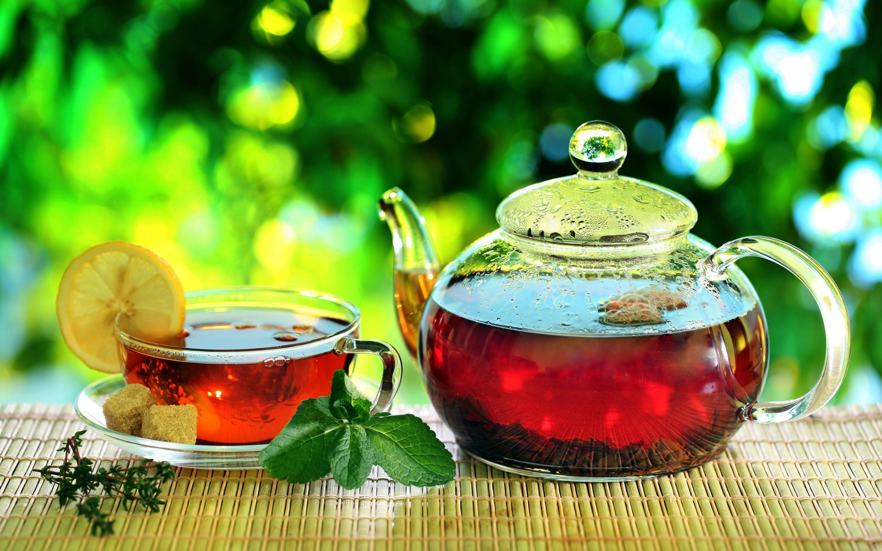 Garden 桌布 壁紙 — 一個玻璃茶壺和杯子盛滿琥珀色茶，放在竹蓆上，旁邊有檸檬片、薄荷和方糖，背景是充滿活力的綠色散景。