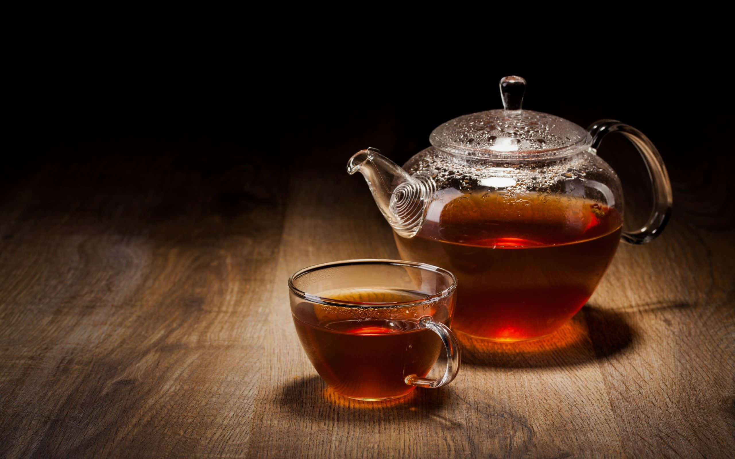tea 桌布 壁紙 — 一個玻璃茶壺和杯子盛滿琥珀色茶，在深色背景下於木質表面上溫暖地發光。