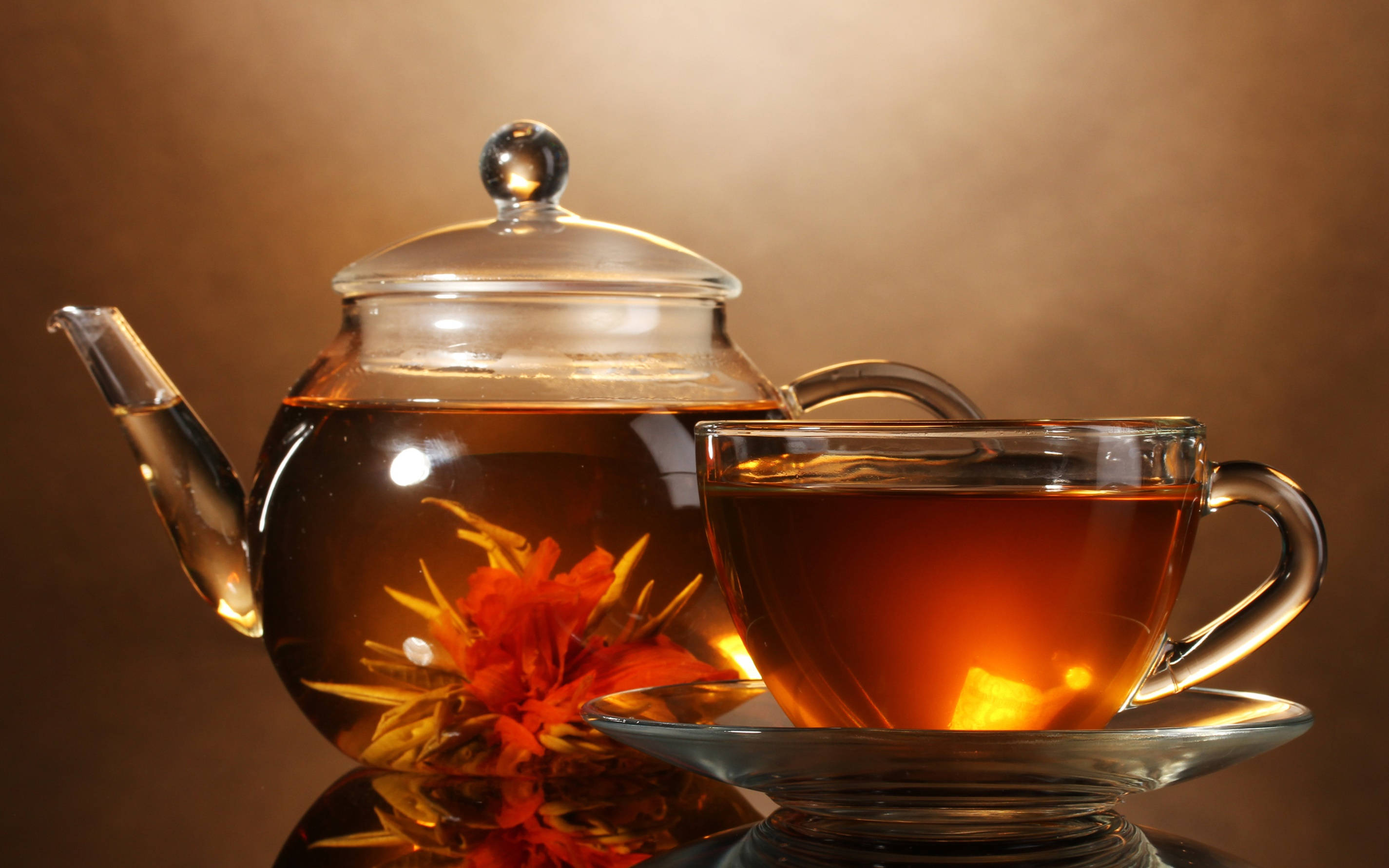 tea 桌布 壁紙 — 一個玻璃茶壺和杯子裝滿琥珀色茶，展示壺內盛開的橙色花朵，背景是溫暖的棕色。