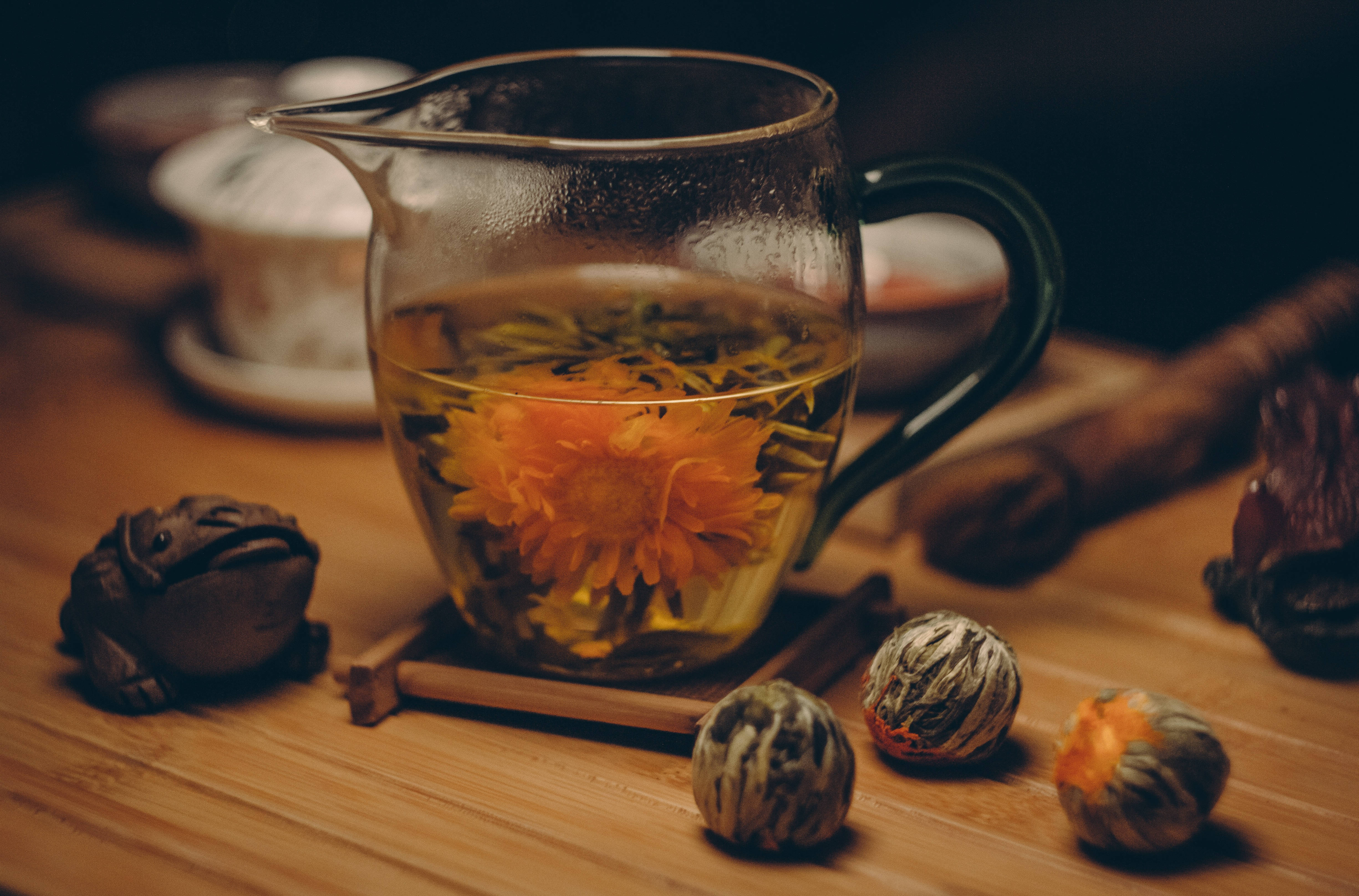 Party 桌布 壁紙 — 一個玻璃茶壺盛載著盛開的橙色菊花茶花，放在溫暖的木桌上，附近有裝飾青蛙公仔和茶球。