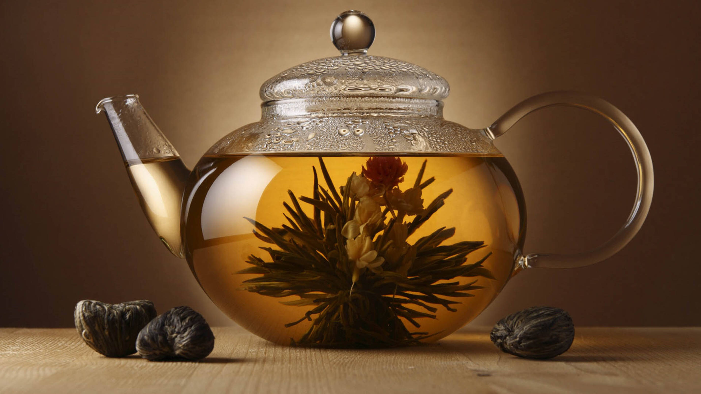 背景 桌布 壁紙 — 一個玻璃茶壺裝滿琥珀色茶和盛開的花蕾，放在木質表面上，附近有乾茶球。