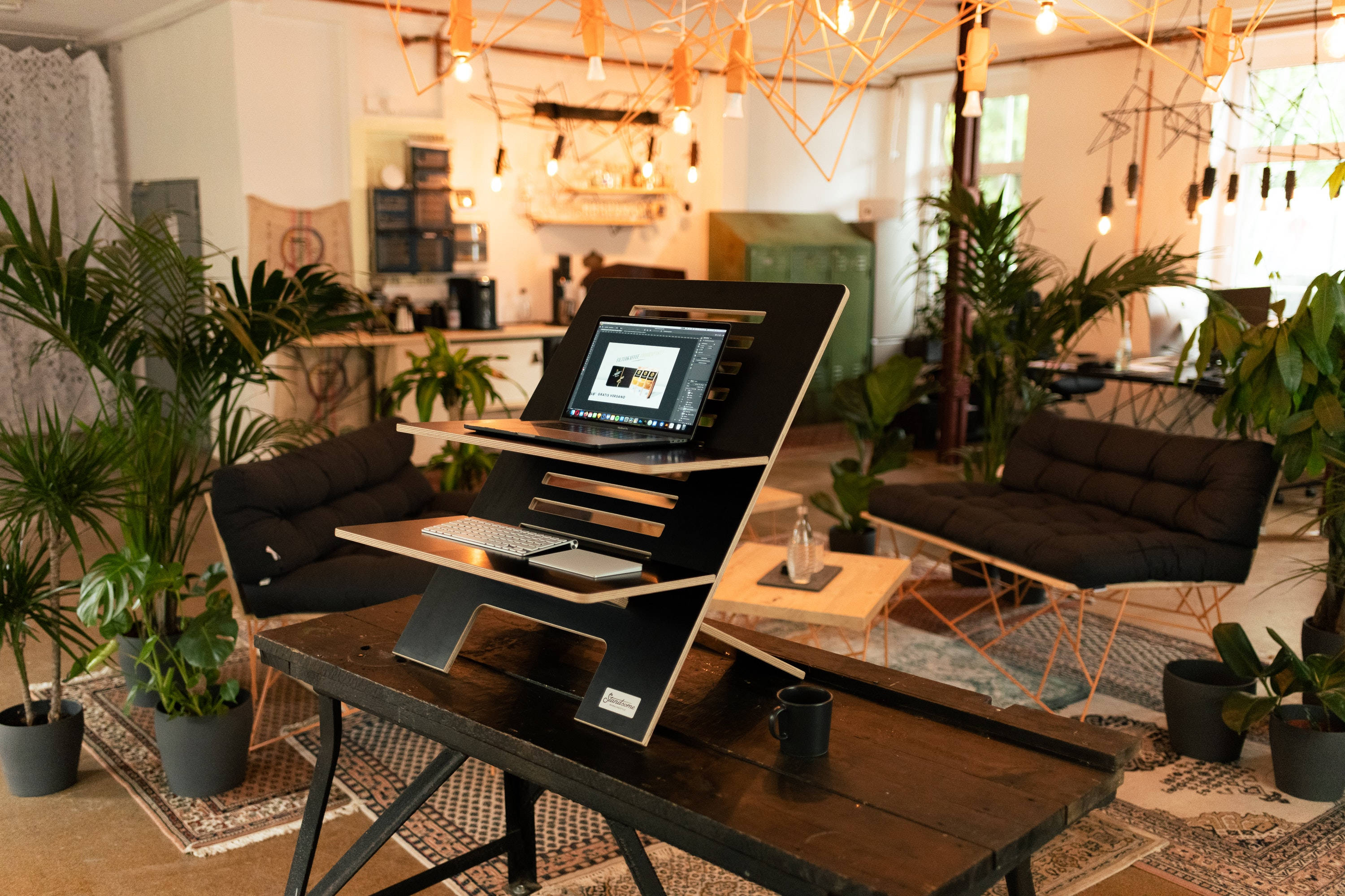 aesthetic tumblr laptop 桌布 壁紙 — 一個現代人體工學書桌設置，配備筆記型電腦和鍵盤，放在黑色可調節支架上，位於舒適且充滿植物的閣樓工作空間。