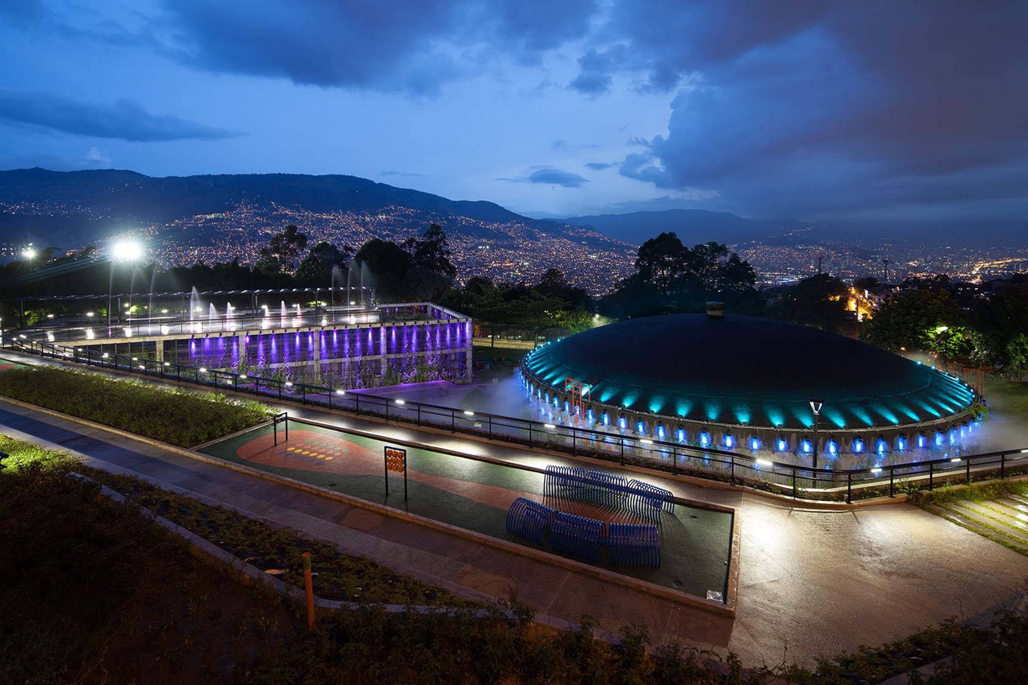 Medellin 桌布 壁紙 — 一個現代化照明公園的黃昏景色，擁有發光的噴泉和一個圓頂結構，俯瞰著廣闊的城市景觀。