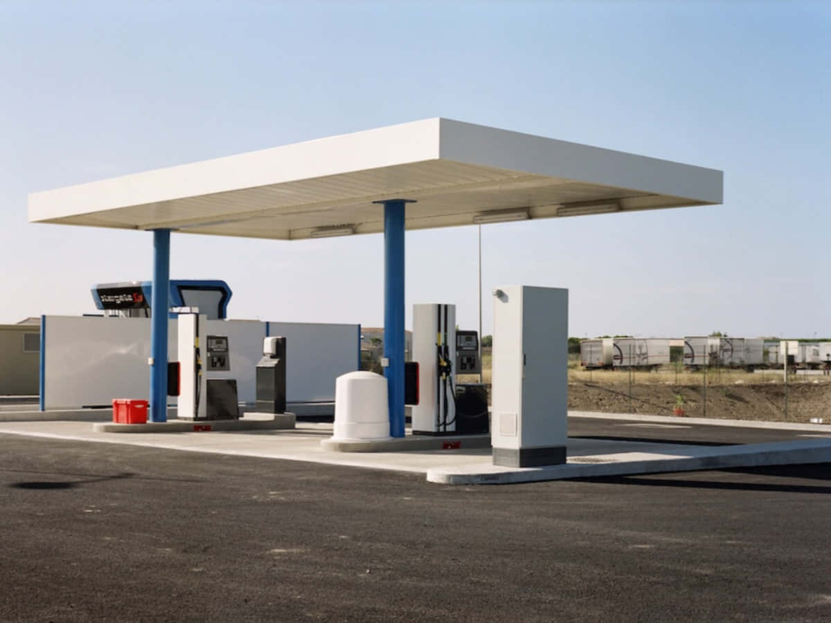 Gas Station 桌布 壁紙 — 一個現代化的加油站，白色頂篷和藍色柱子，矗立在晴朗的藍天下，空蕩蕩的瀝青地面上。