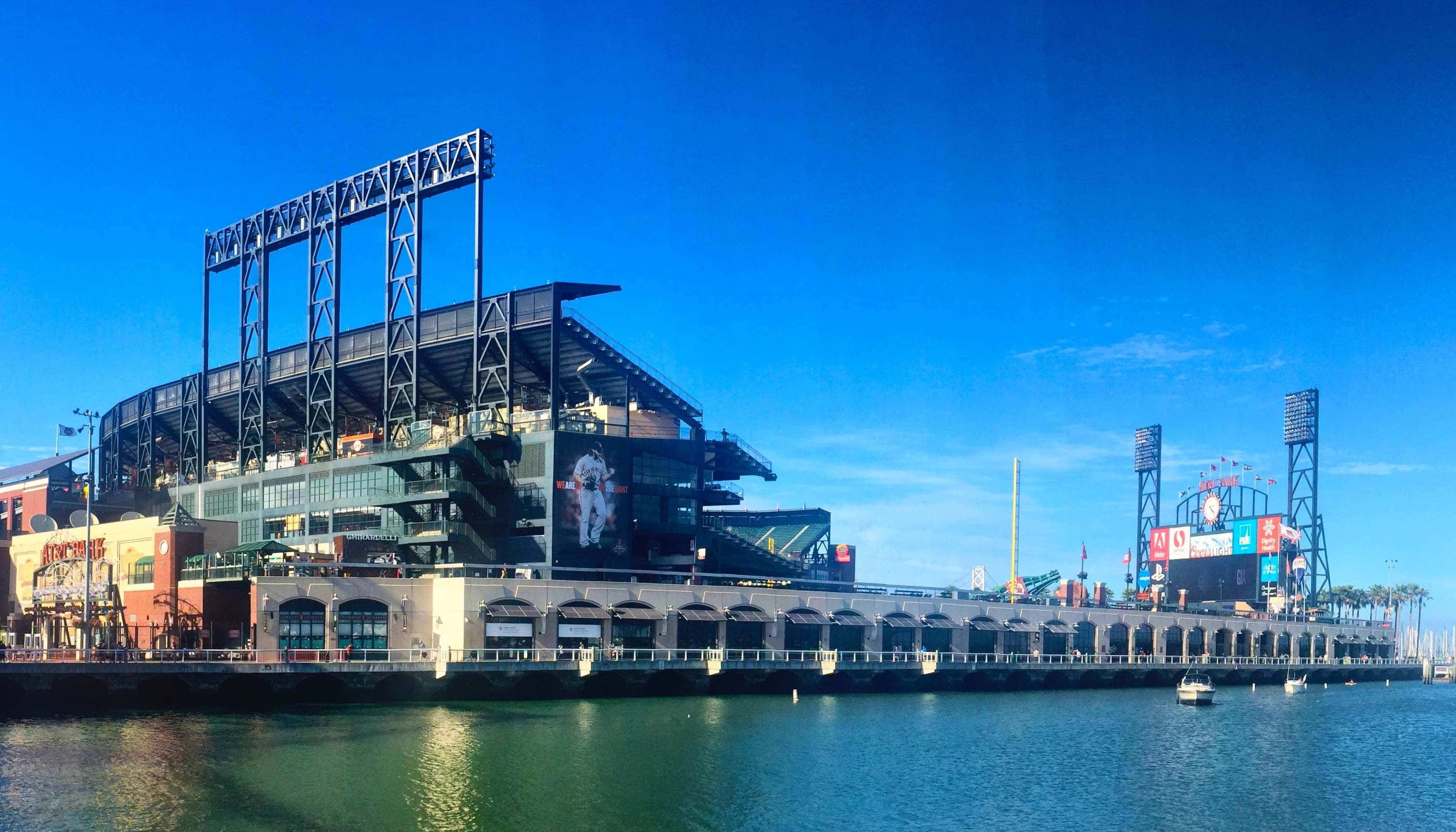 Baseball Stadium 桌布 壁紙 — 一個現代化的棒球場，採用鋼結構框架和玻璃外牆，矗立在平靜的水邊，天空清澈湛藍。