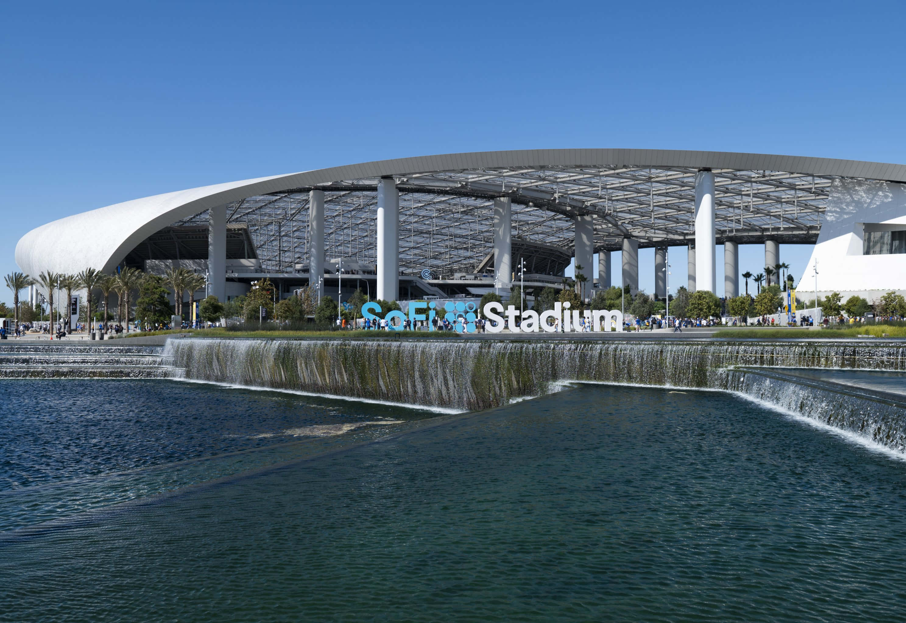 Sofi Stadium 桌布 壁紙 — 一個現代化的體育場，擁有彎曲的白色屋頂和層疊的水景，在晴朗的藍天下。