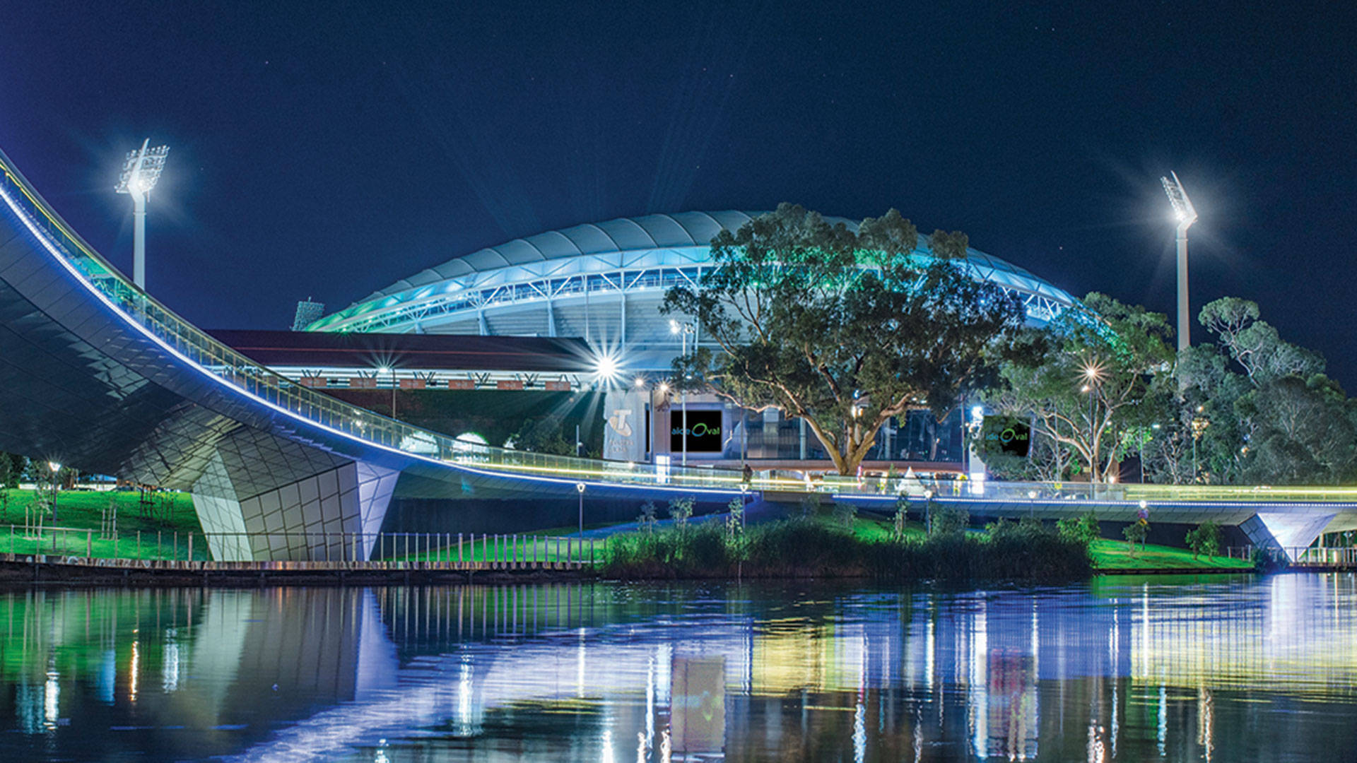 Adelaide 桌布 壁紙 — 一個現代化的體育館和彎曲的橋樑在夜晚發出藍光，它們的倒影在星空下平靜的水面上閃爍。