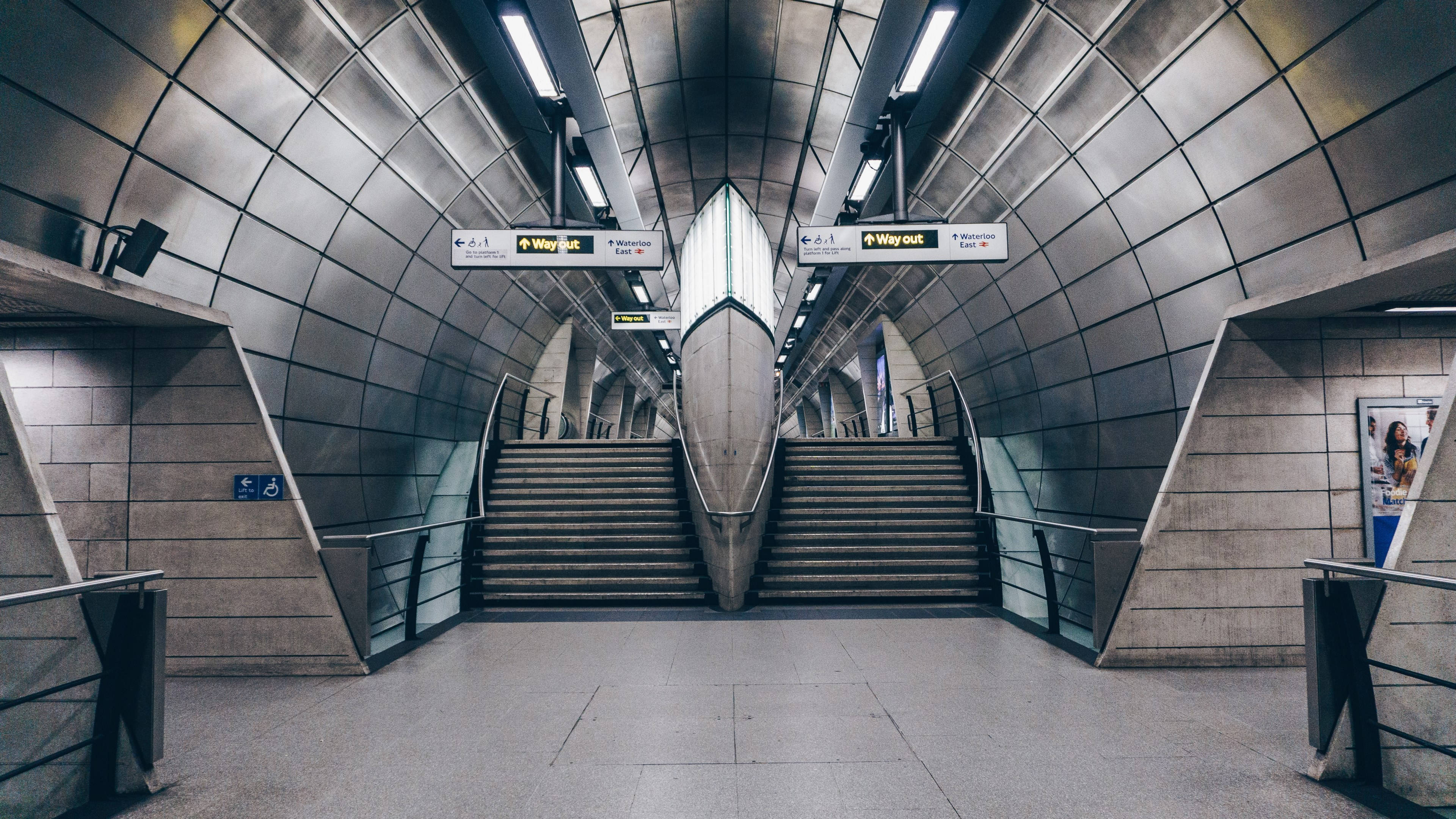 train tunnel 桌布 壁紙 — 一個現代地鐵站的對稱景觀，彎曲的金屬牆壁和雙樓梯在明亮的螢光燈下向上延伸。