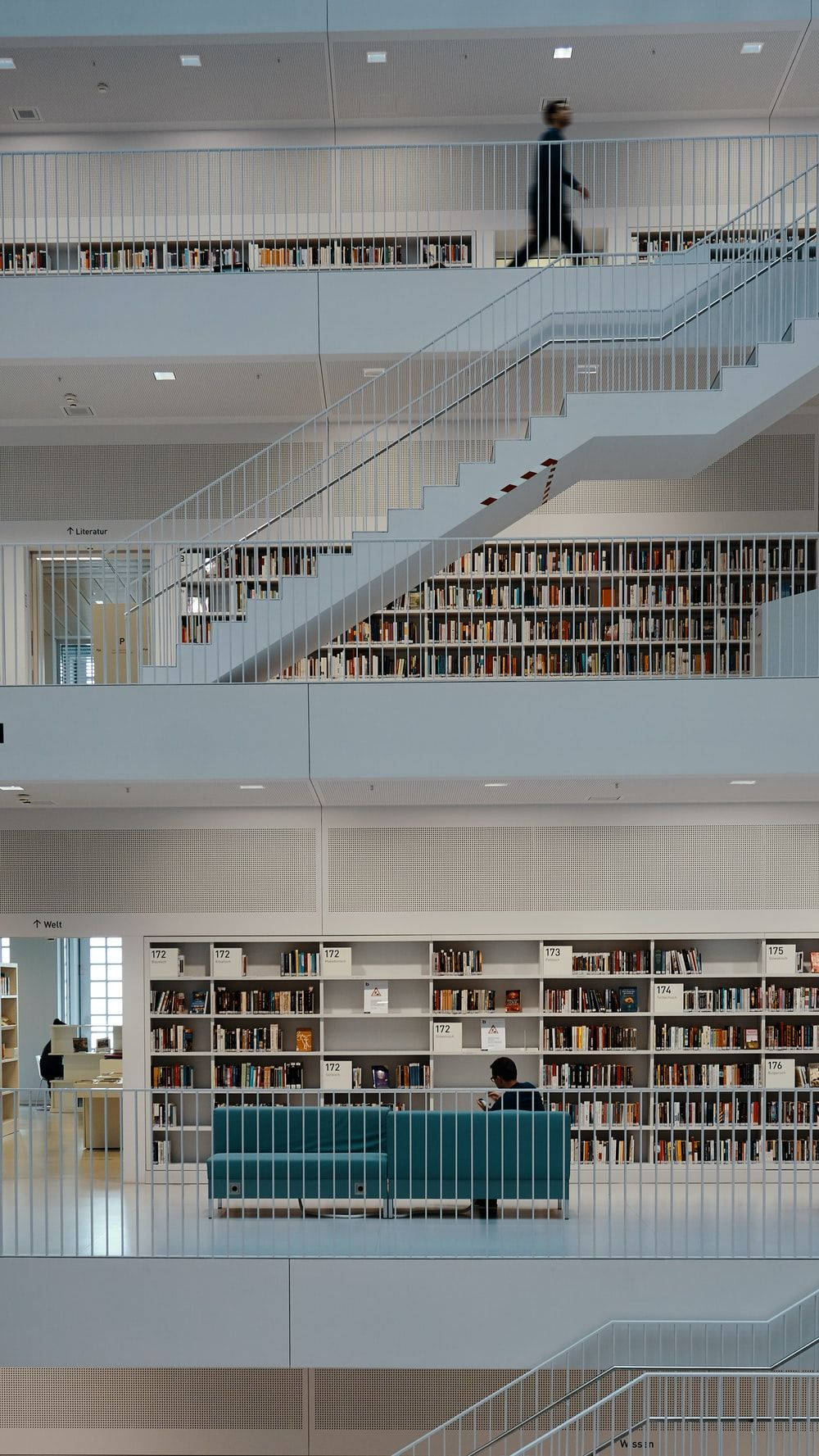 Modern 桌布 壁紙 — 一個現代多層圖書館，設有白色樓梯和裝滿書的書架，還有一個人在青綠色沙發上閱讀。
