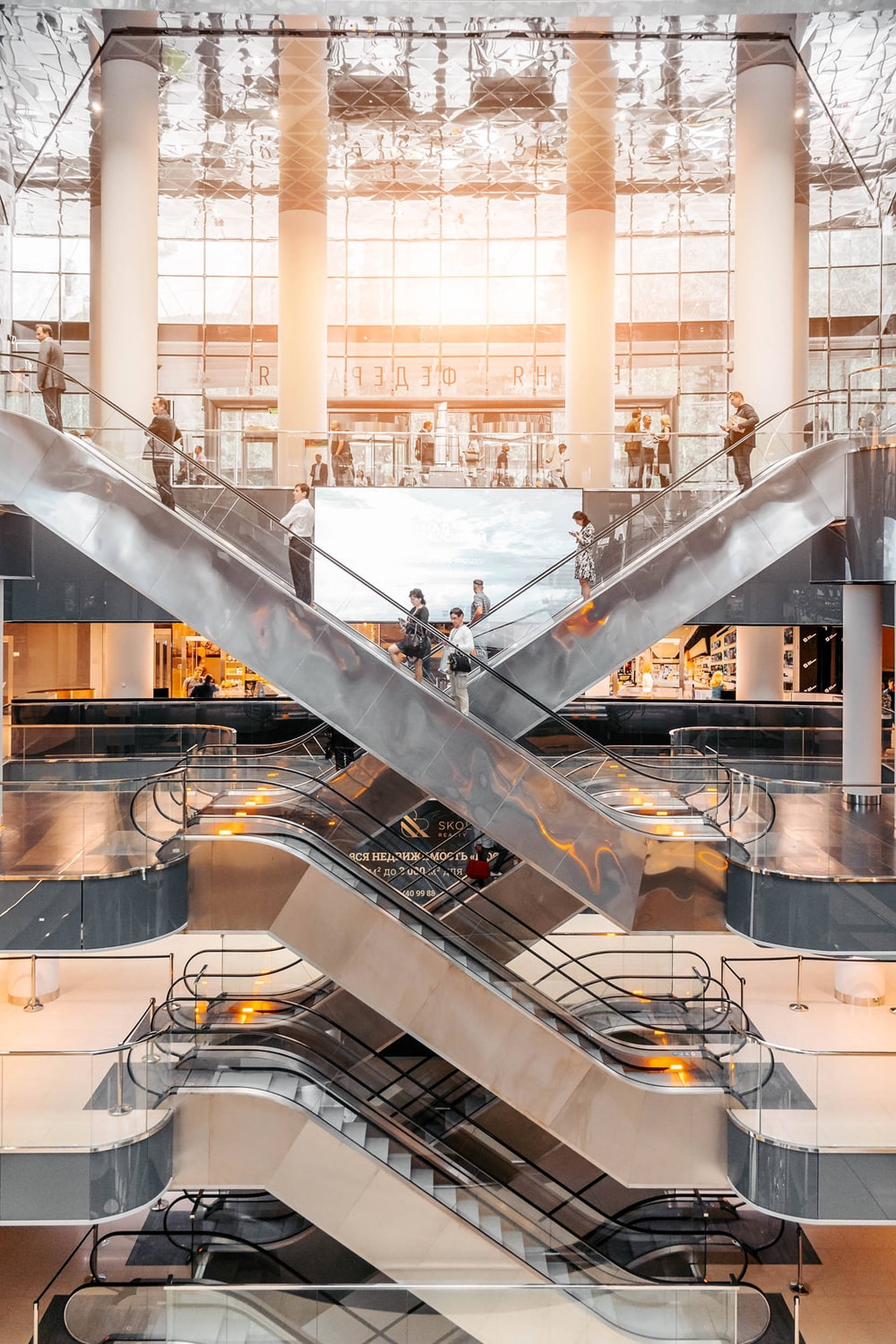 Mall 桌布 壁紙 — 一個現代大型購物中心內部，設有交錯的自動扶梯，人們在玻璃天花板下穿梭於各樓層之間。