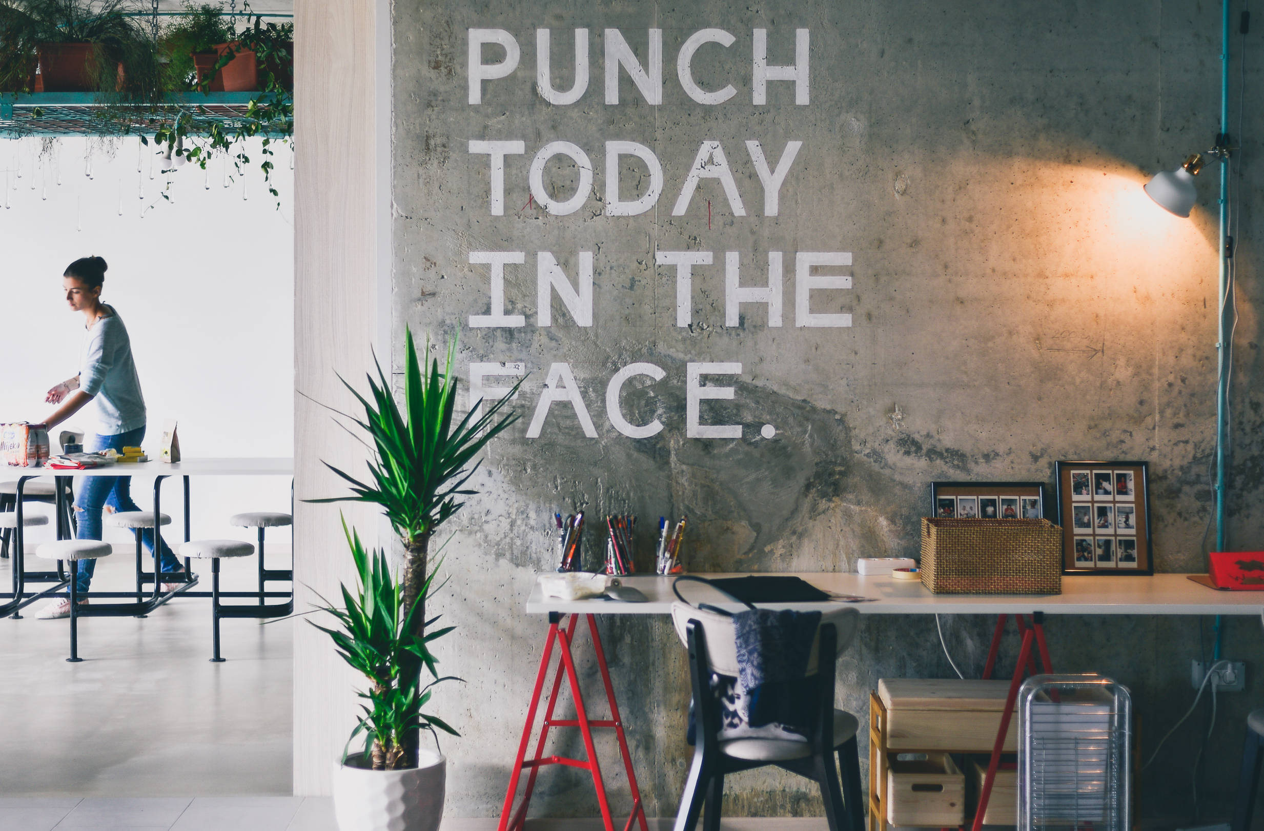 激勵 桌布 壁紙 — 一個現代工作空間，混凝土牆上印有粗體文字「punch today in the face」，附近有一人在桌子上工作。