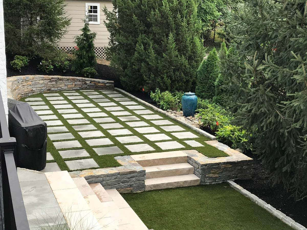 Landscaping 桌布 壁紙 — 一個現代後院露台，採用石鋪網格圖案，周圍環繞著茂密的綠植和常綠樹木。