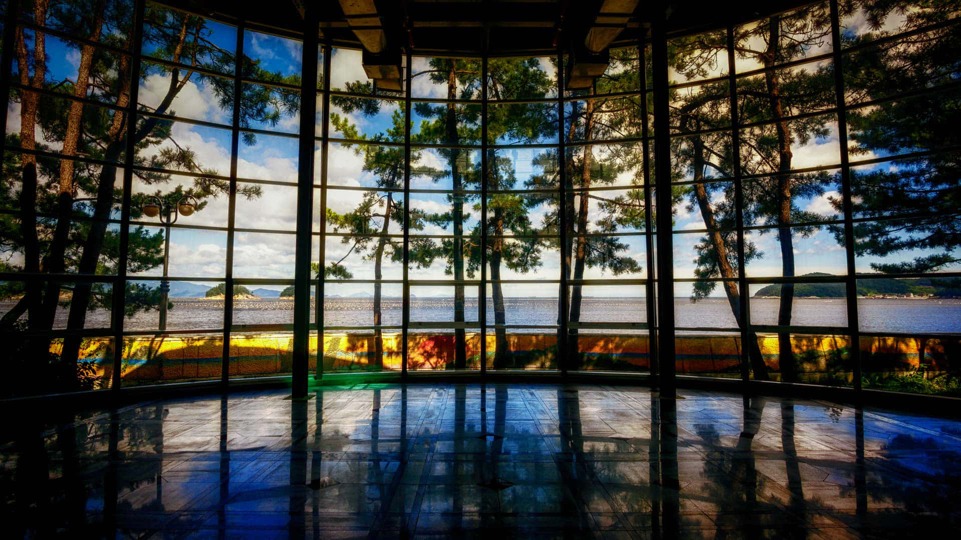Museums 桌布 壁紙 — 一個現代玻璃牆房間反射出寧靜的湖泊和松樹，在明亮的藍天和散落的雲朵下。