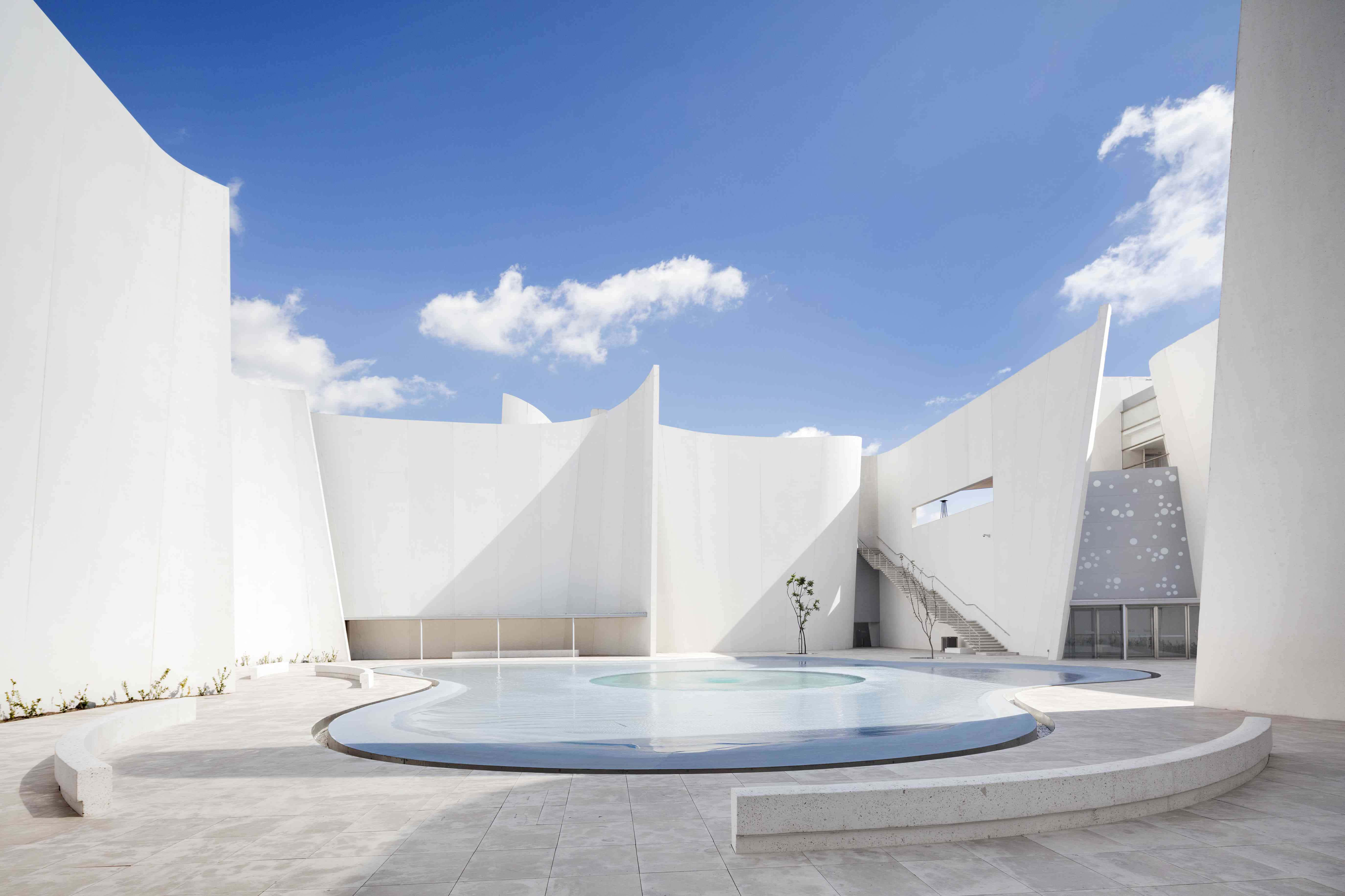 Minimalist Architecture 桌布 壁紙 — 一個現代白色建築庭院，設有彎曲泳池，上方是帶有散雲的明亮藍天。