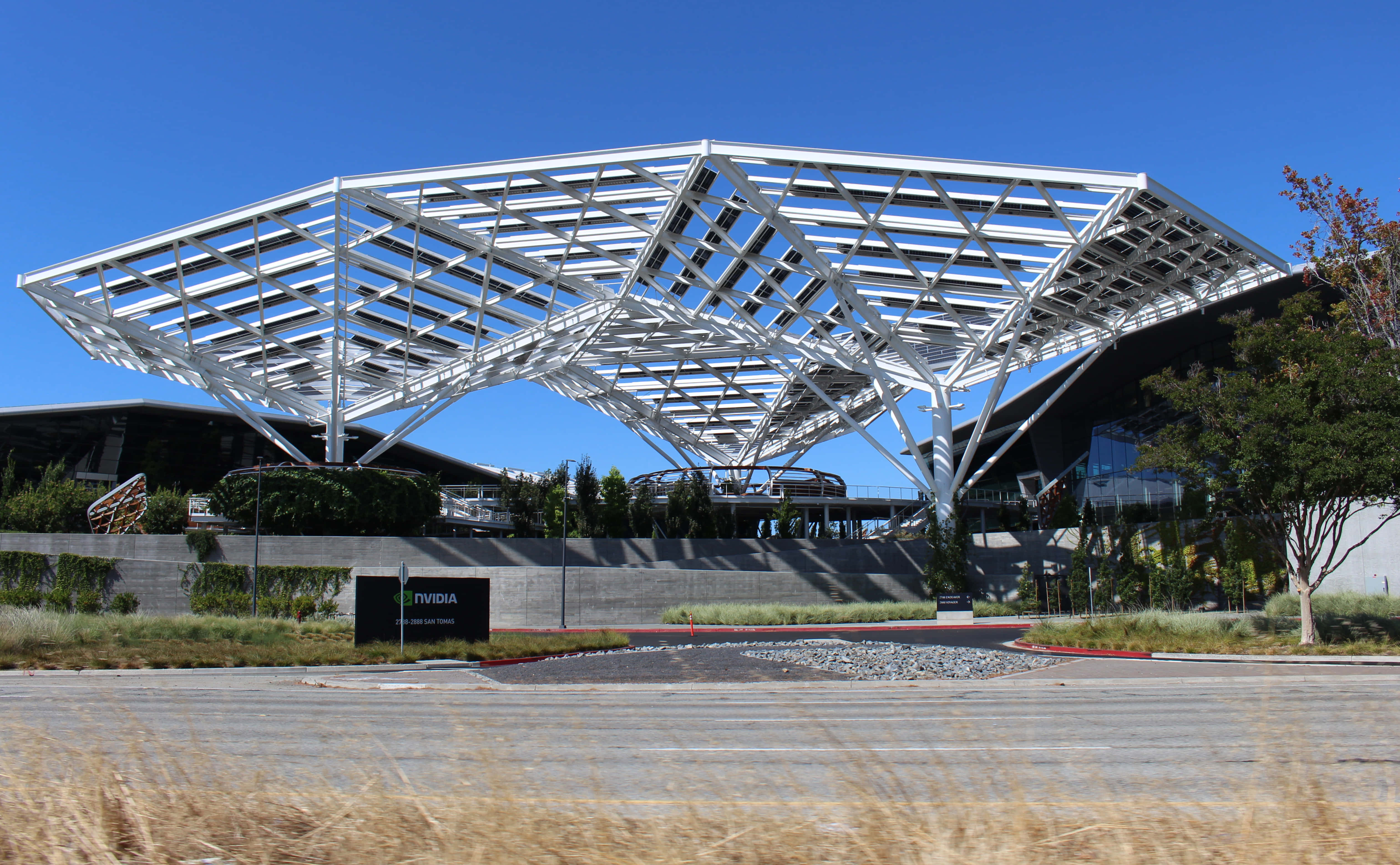 nvidia 桌布 壁紙 — 一個現代白色鋼格柵天篷拱起在鋪砌的廣場上，廣場上有乾草和Nvidia標誌，在晴朗的藍天下。