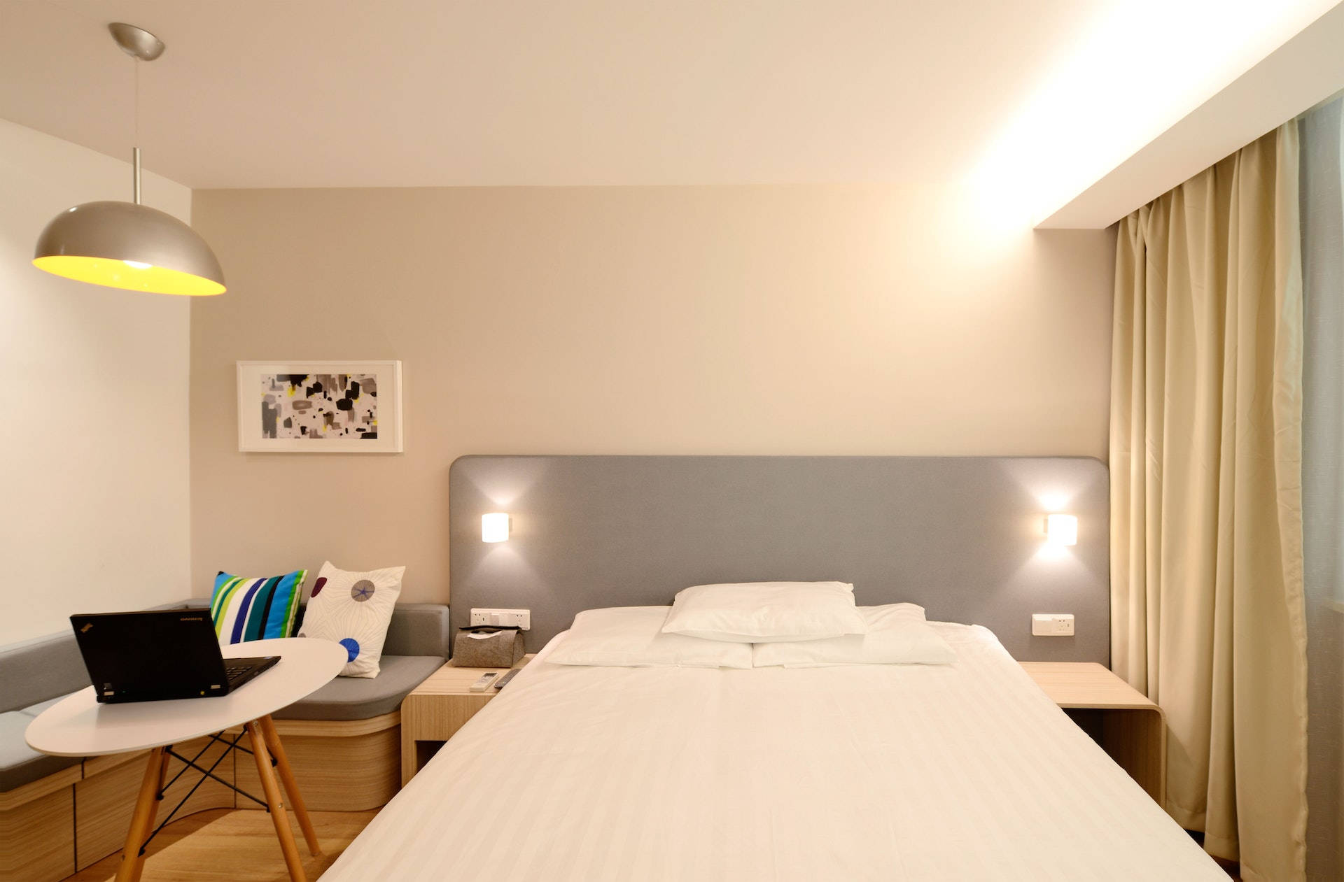 房屋 桌布 壁紙 — 一個現代簡約風格的臥室，配有整齊鋪好的床、溫暖的燈光，以及內置長凳旁圓桌上的手提電腦。