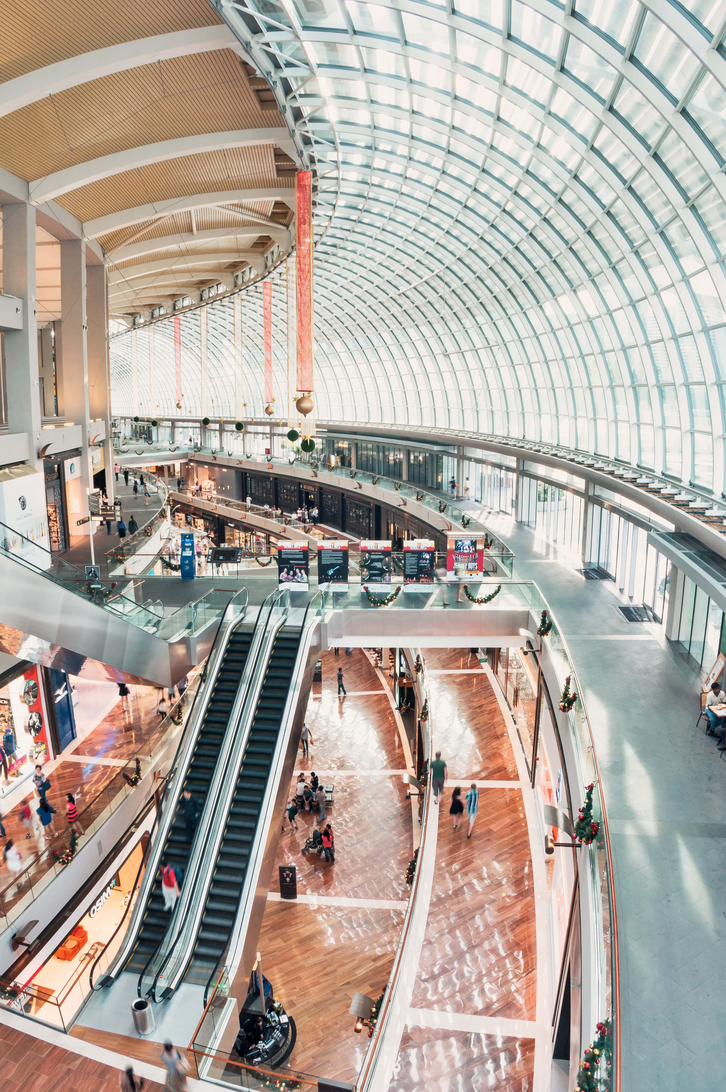 Mall 桌布 壁紙 — 一個現代購物中心的高角度視圖，設有弧形玻璃天花板、自動扶梯和購物者在木地板上行走。