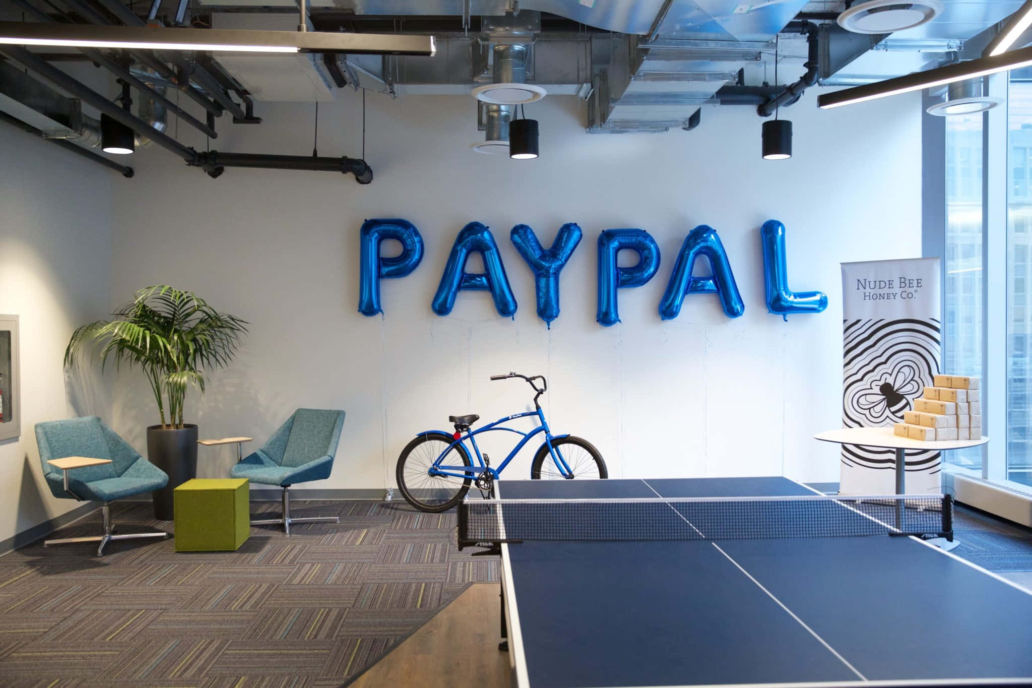 PayPal 桌布 壁紙 — 一個現代辦公室休息室，設有藍色PayPal氣球標誌、腳踏車和乒乓球桌，下方是裸露的天花燈。