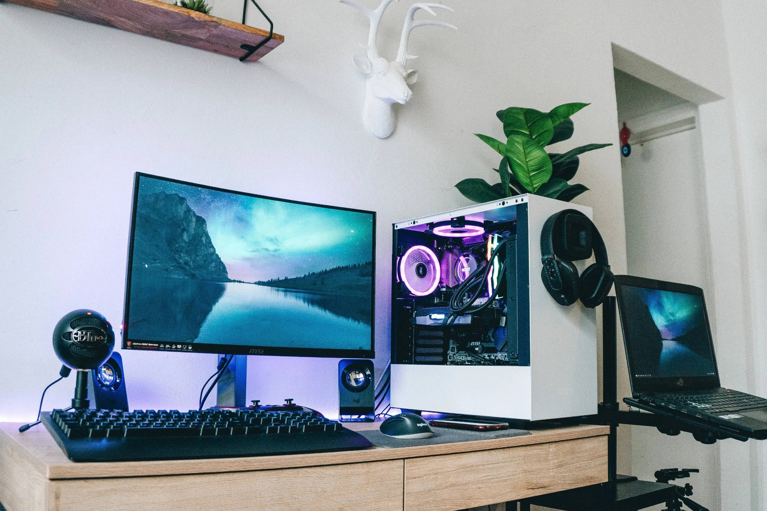 Gaming Setup 桌布 壁紙 — 一個現代遊戲設置，配備曲面顯示器，顯示暮色中寧靜的湖泊，以及帶有發光紫色內部的白色PC。