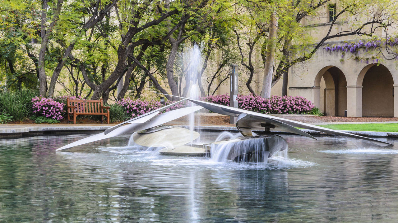 Pond 桌布 壁紙 — 一個現代金屬噴泉，流水潺潺，坐落於寧靜的花園中，周圍環繞著樹木和盛開的粉紅色花朵。