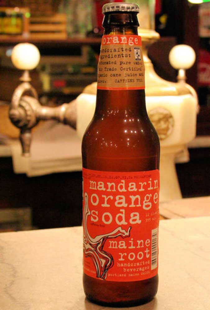 Drink 桌布 壁紙 — 一個琥珀色玻璃瓶裝的柑橘橙味汽水，帶有鮮明的紅色標籤，放在一個帶有復古水龍頭的酒吧櫃檯上，光線柔和溫暖。