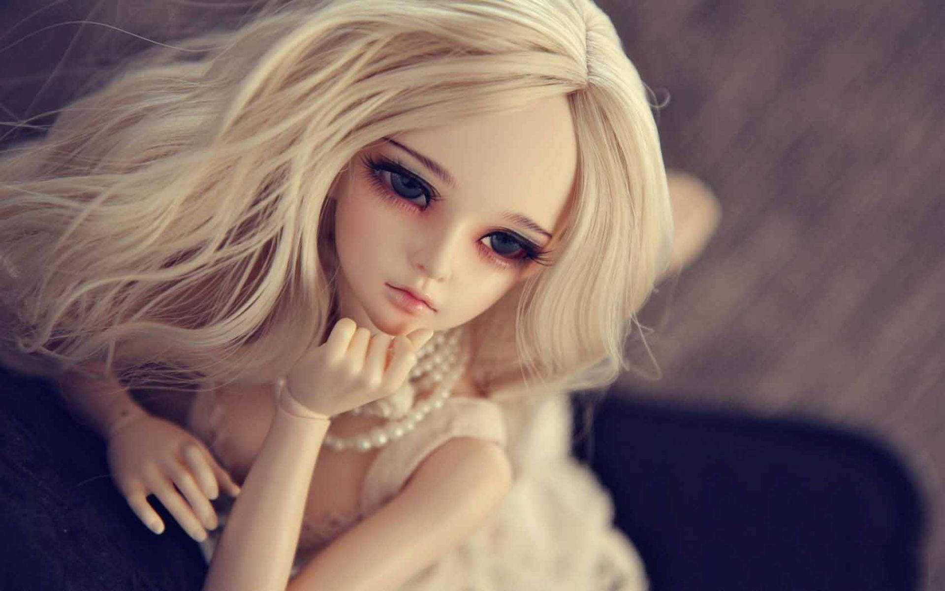 Doll 桌布 壁紙 — 一個瓷娃娃，擁有飄逸的金色長髮和一雙藍色大眼睛，戴著珍珠，在柔和的模糊背景下若有所思地休息。